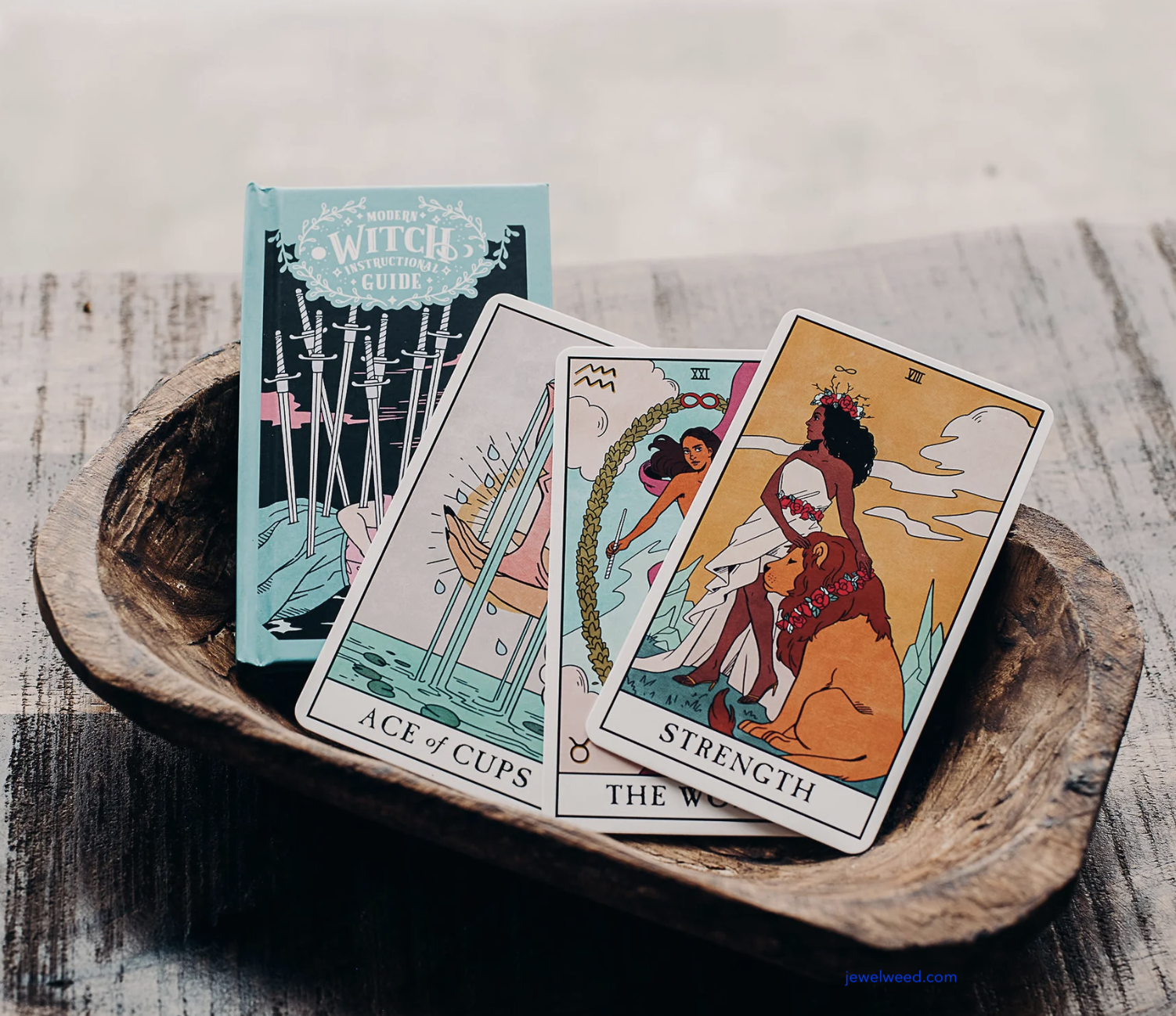 Tarot cards laid out in a wooden bowl, including the Ace of Cups, The World, and Strength, with a guidebook behind.
