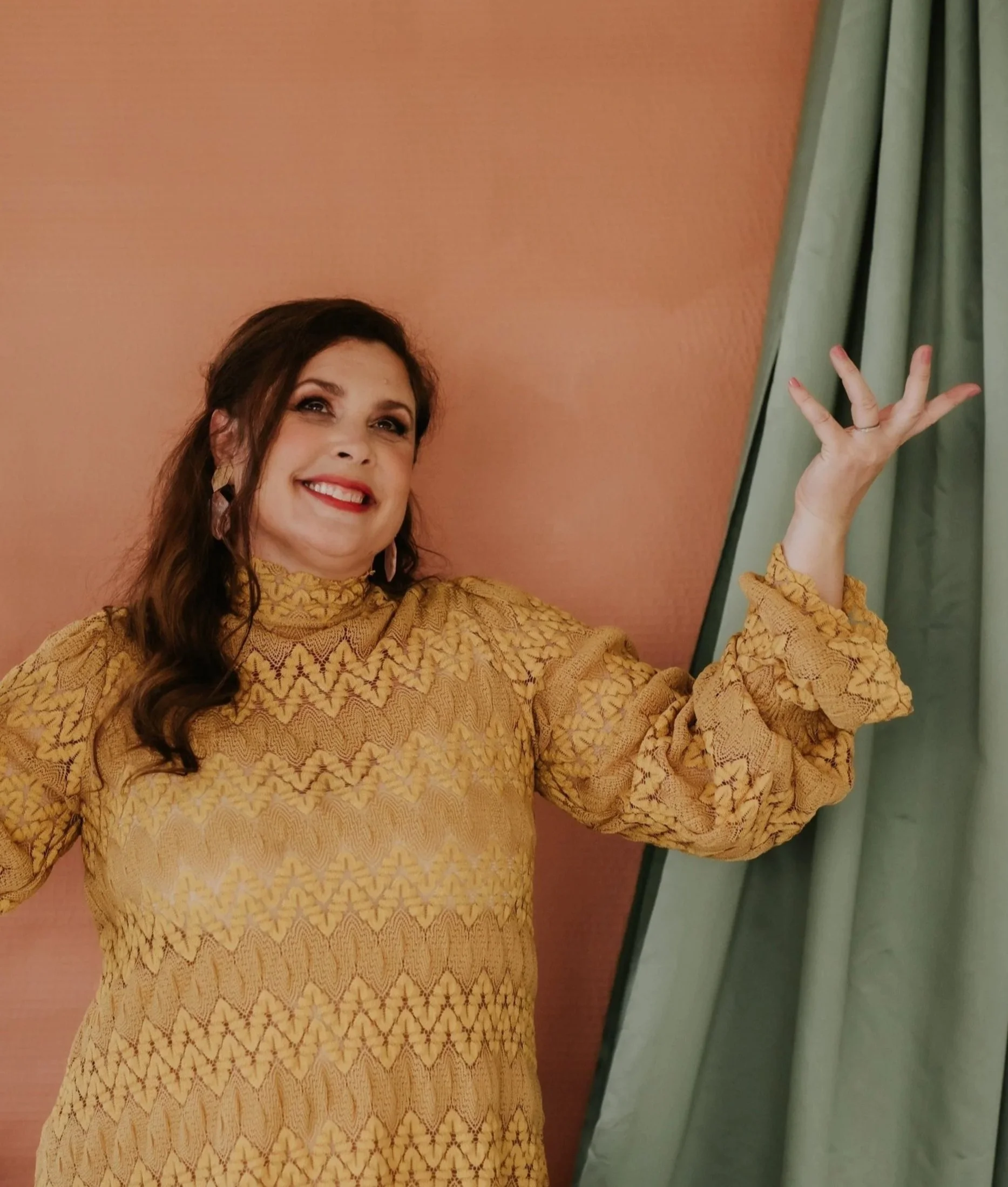 Woman with brown hair and makeup smiling, wearing a mustard yellow patterned dress with puffed sleeves, standing in front of a pink wall and green curtains, gesturing with her right hand.