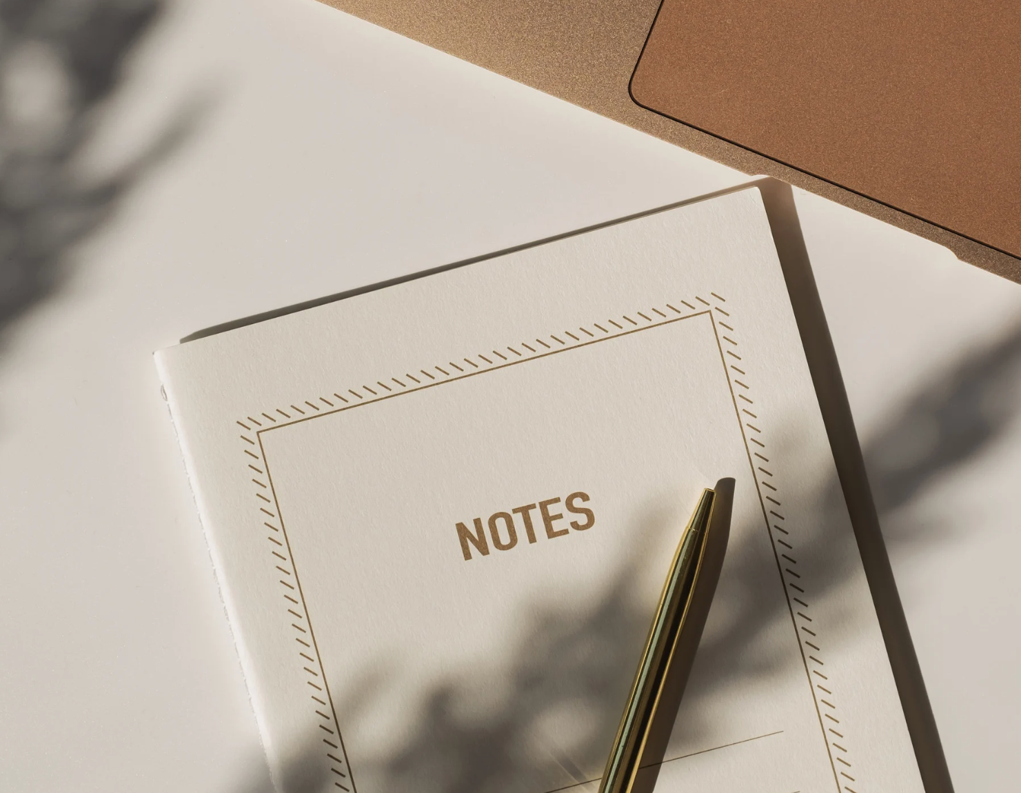 A white notebook with 'NOTES' written on the cover, a gold pen resting on it, and a pinkish-brown box in the background, all on a light surface.