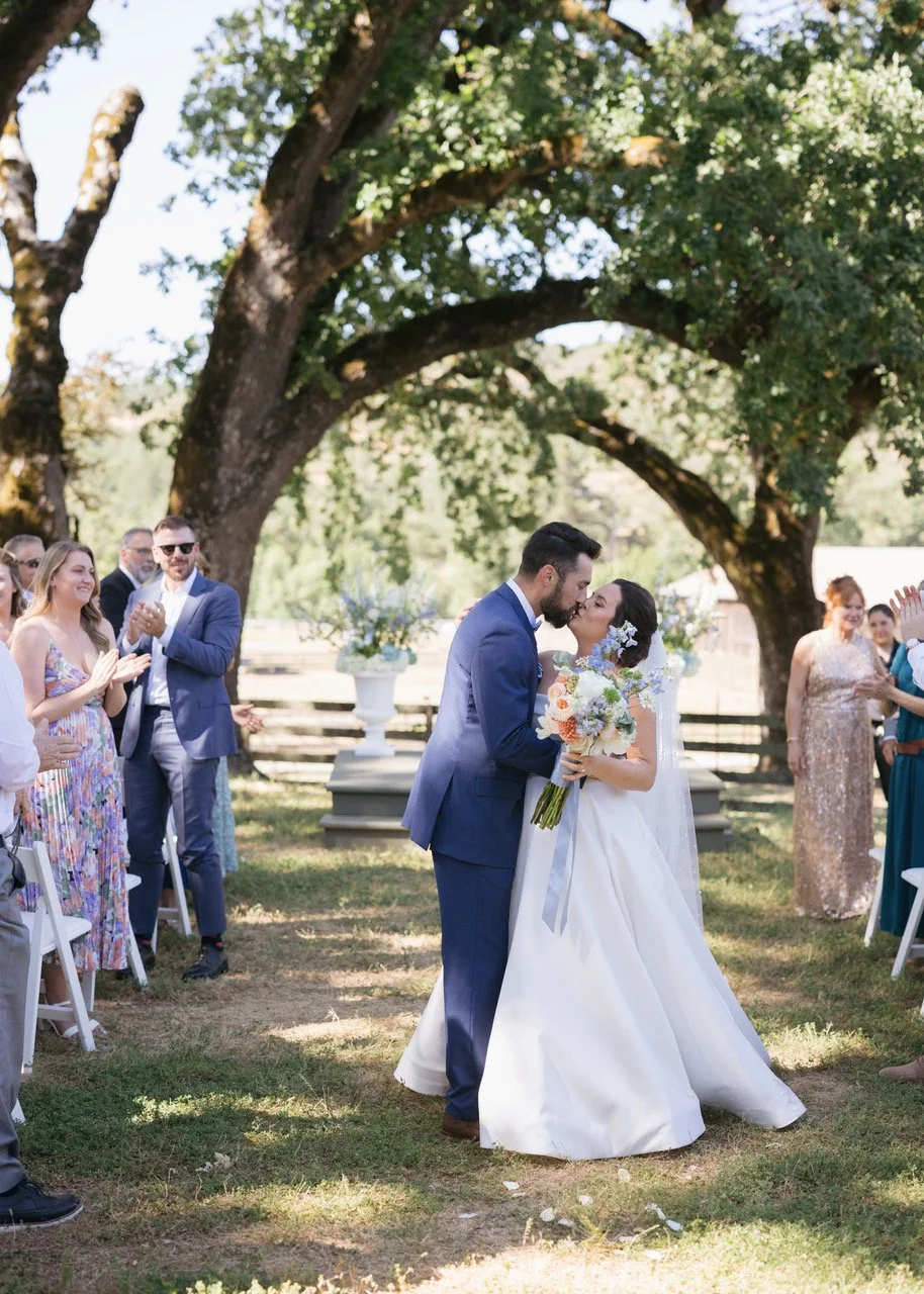 A wedding couple sharing a kiss outdoors under a large tree, with friends clapping and smiling around them.