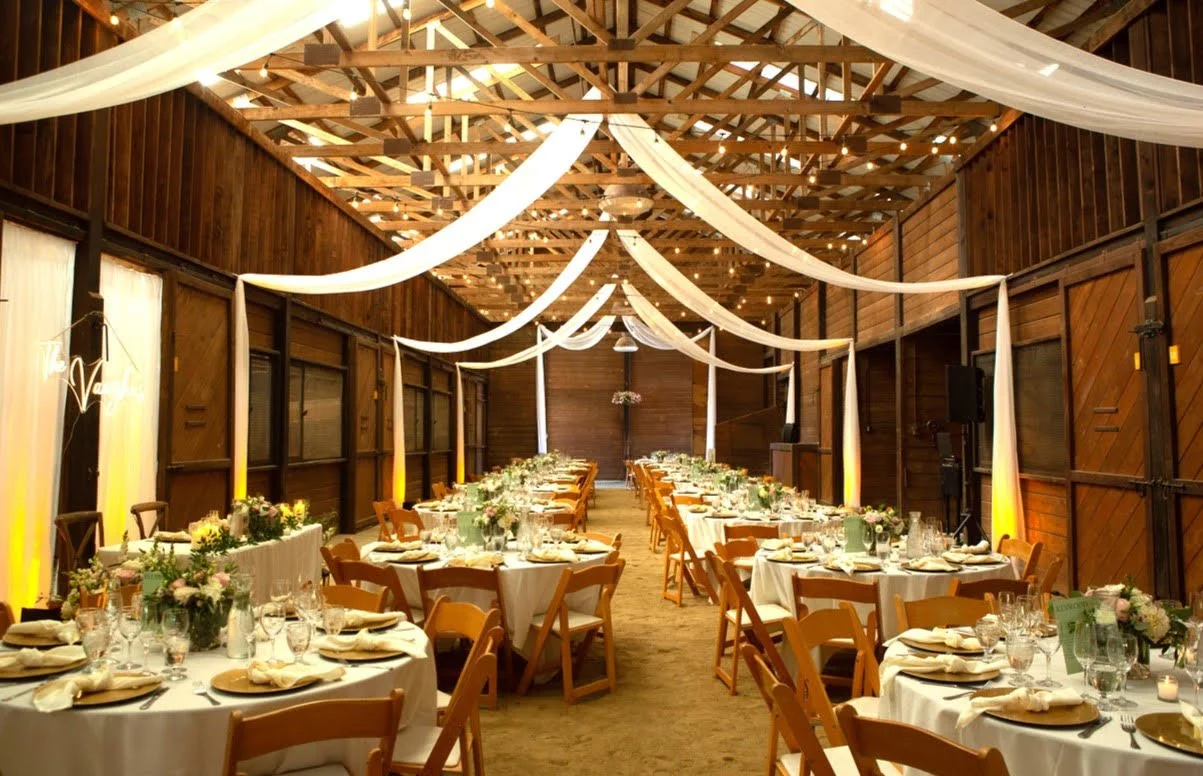 A beautifully decorated wedding reception hall with round tables covered with white tablecloths, floral centerpieces, and elegant place settings. White fabric drapes are hanging from the wooden ceiling beams, and warm lighting is illuminating the woo