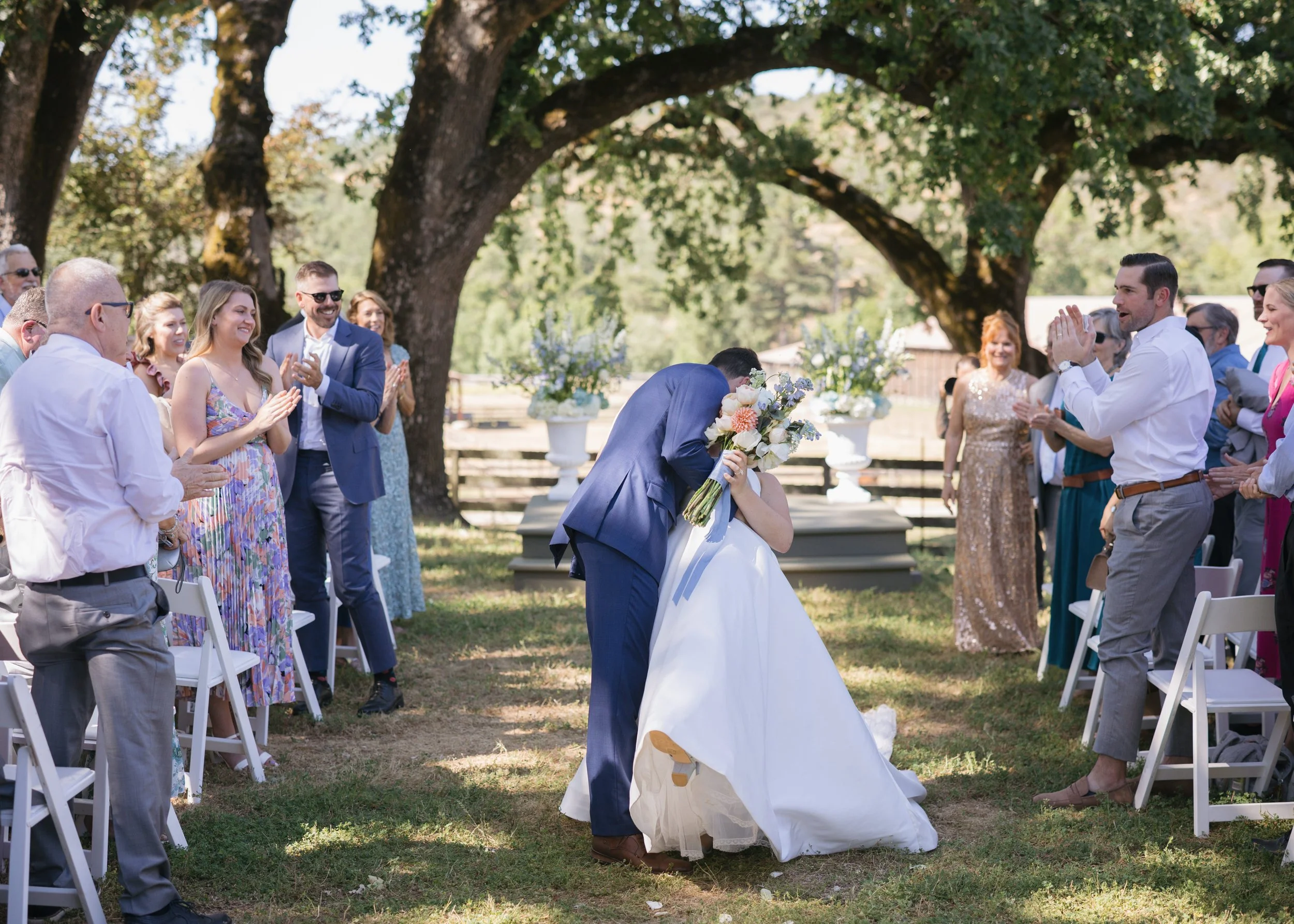 A newlywed couple shares a kiss during their outdoor wedding ceremony, surrounded by friends and family on a sunny day under large trees.