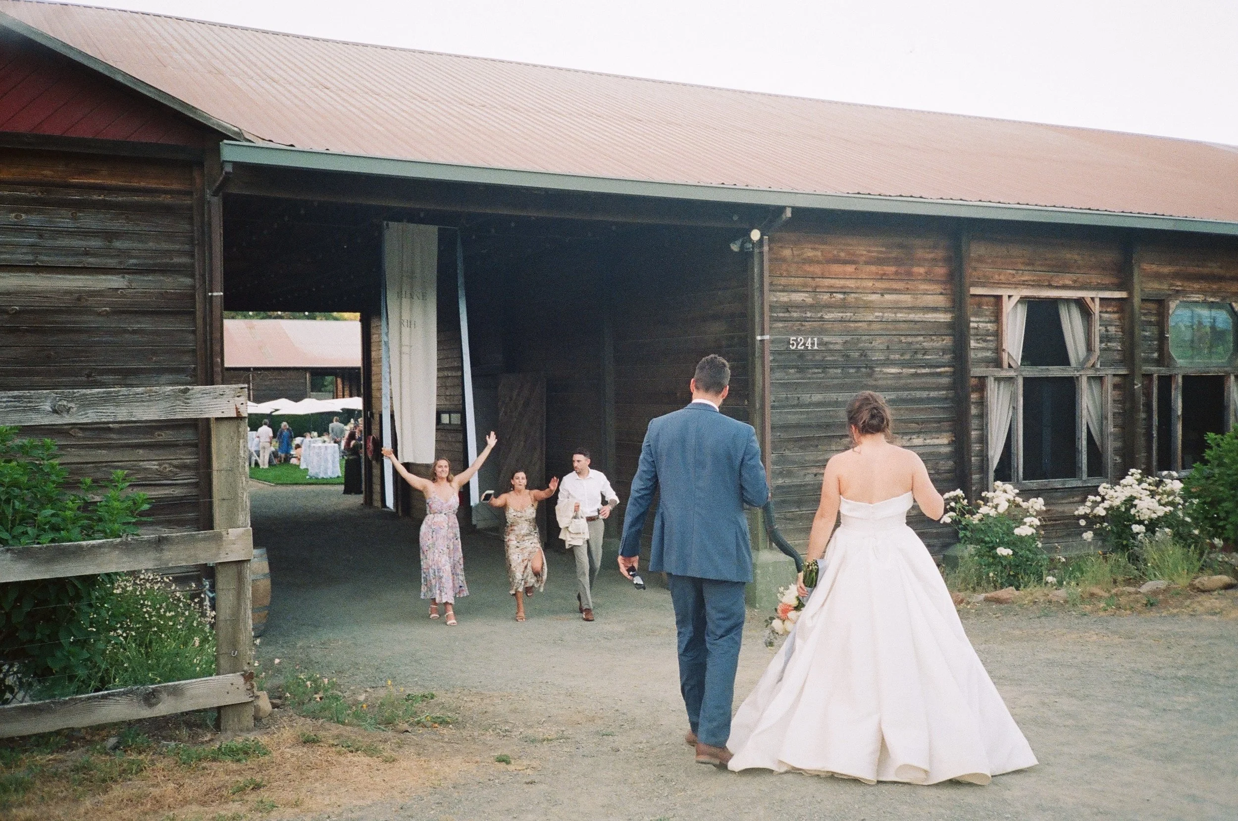 Bride and groom entering a rustic barn at a wedding, with guests cheerfully greeting them from outside.