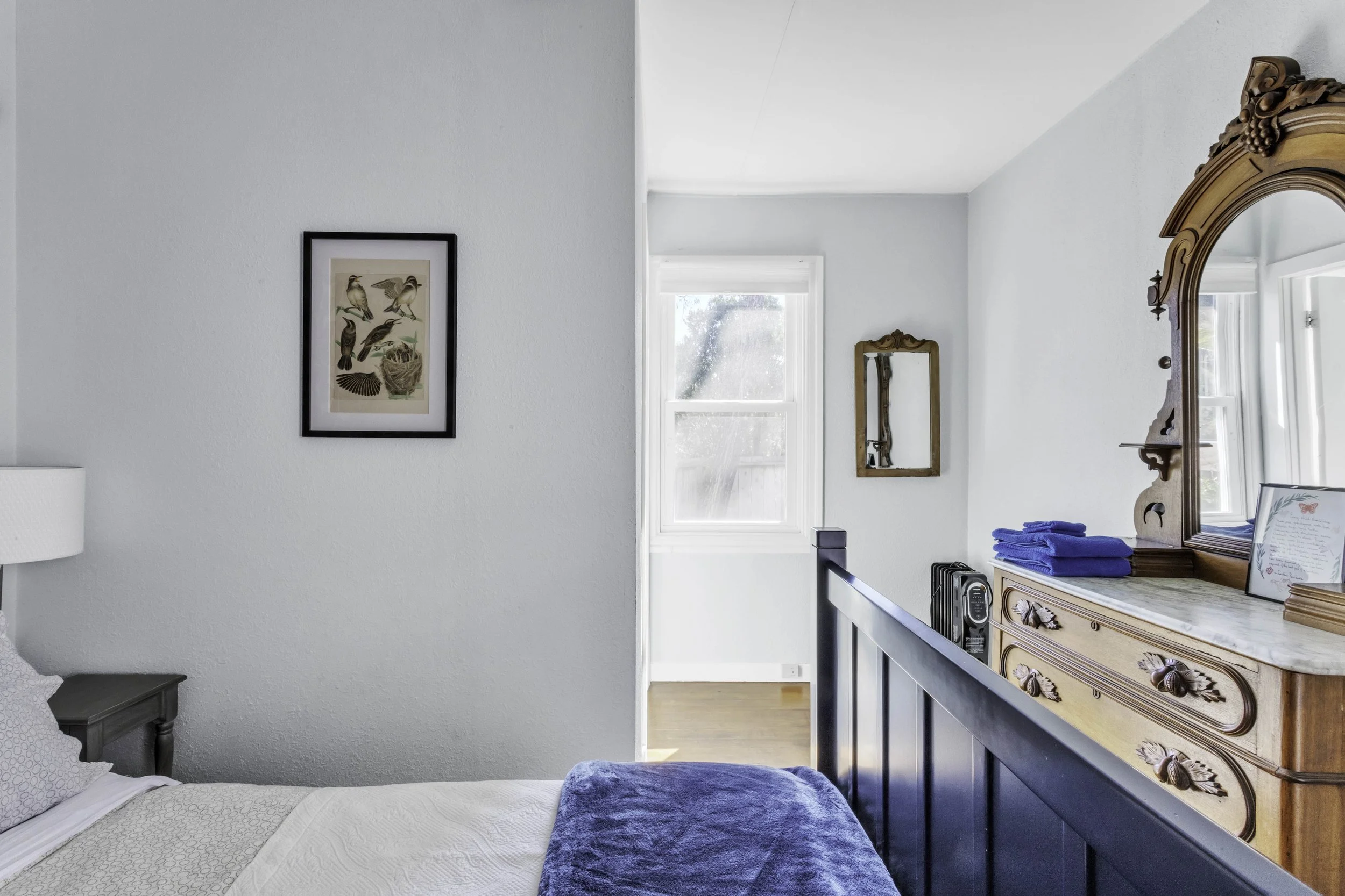 A bedroom with a bed, a lamp, artwork on the walls, a window, and a wooden dresser with a mirror, some blue towels, and framed items on top.