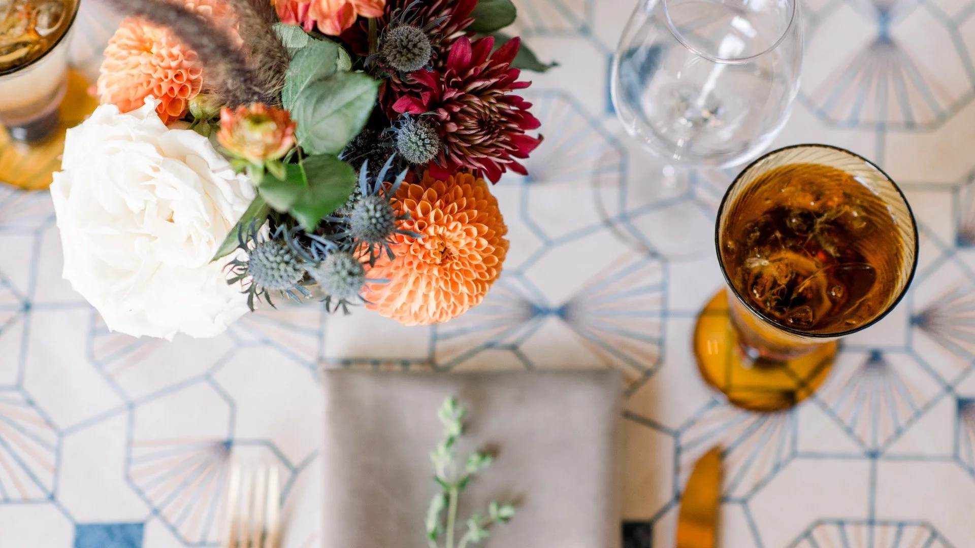 A top-down view of a table setting featuring a floral centerpiece with white, orange, red, and purple flowers, and glasses of drinks, one with dark liquid and ice cubes. The table has a geometric patterned tablecloth.