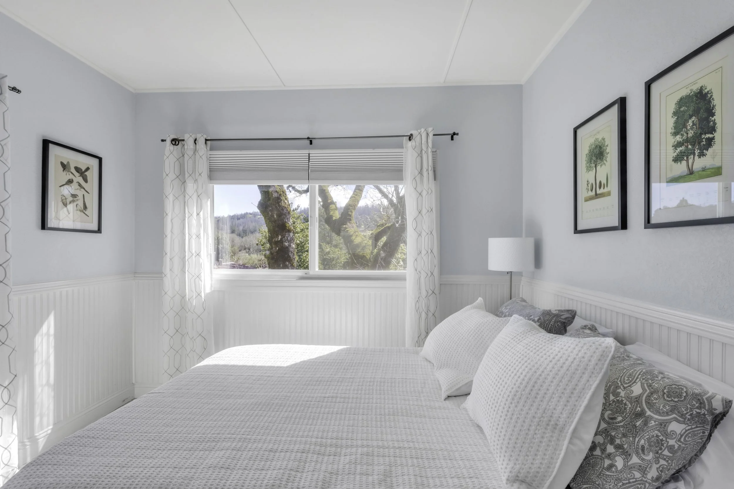 A bright bedroom with a large window showing trees outside. The bed has white bedding and several pillows, with wall art featuring trees and birds above it.