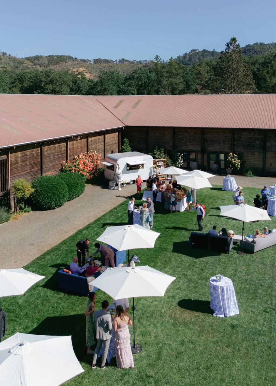 Outdoor gathering with guests socializing under white umbrellas on a lawn, near a wooden building with a metal roof, with hills and trees in the background.