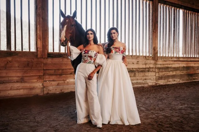 Two women in white dresses with floral tops standing next to a black horse inside a wooden stable.