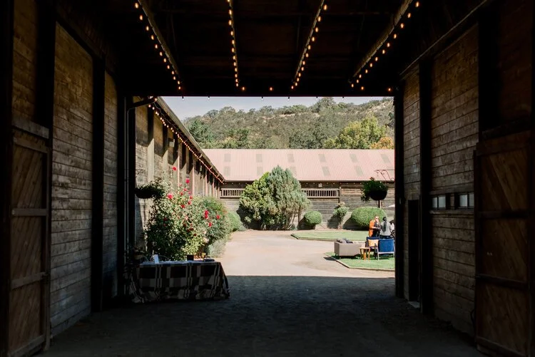 View from inside a rustic barn, looking out onto a courtyard with trees and bushes, outdoor seating, and a couple of people sitting and chatting; string lights hanging from the barn ceiling.