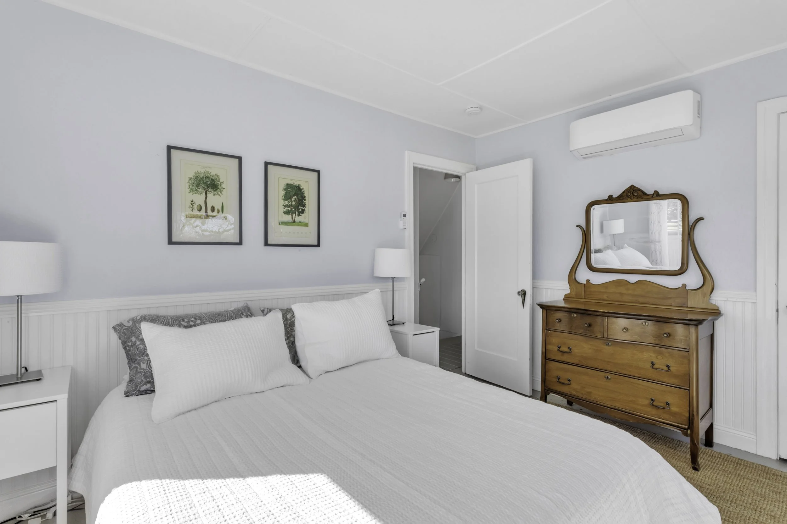 A bedroom with a white bed, white pillows, two white nightstands with lamps, framed artwork of trees on the wall, an antique wooden dresser with a mirror, and an air conditioning unit on the wall.