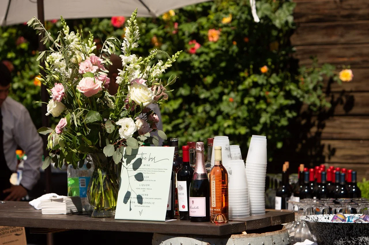 A table with flower arrangement, bottles of wine, plastic cups, and a sign at an outdoor event.