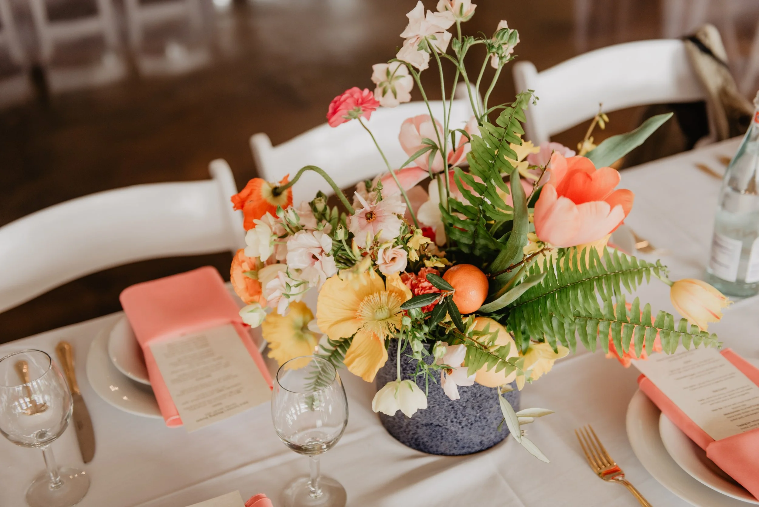 A table centerpiece with a variety of colorful flowers including pink tulips, white and pink sweet peas, and ferns in a dark textured vase, set on a white tablecloth with pink napkins, wine glasses, and place settings.