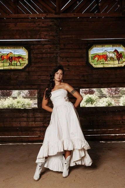 A woman in a white strapless ruffled dress standing inside a barn with dark wood walls, with two small windows behind her showing painted horses.
