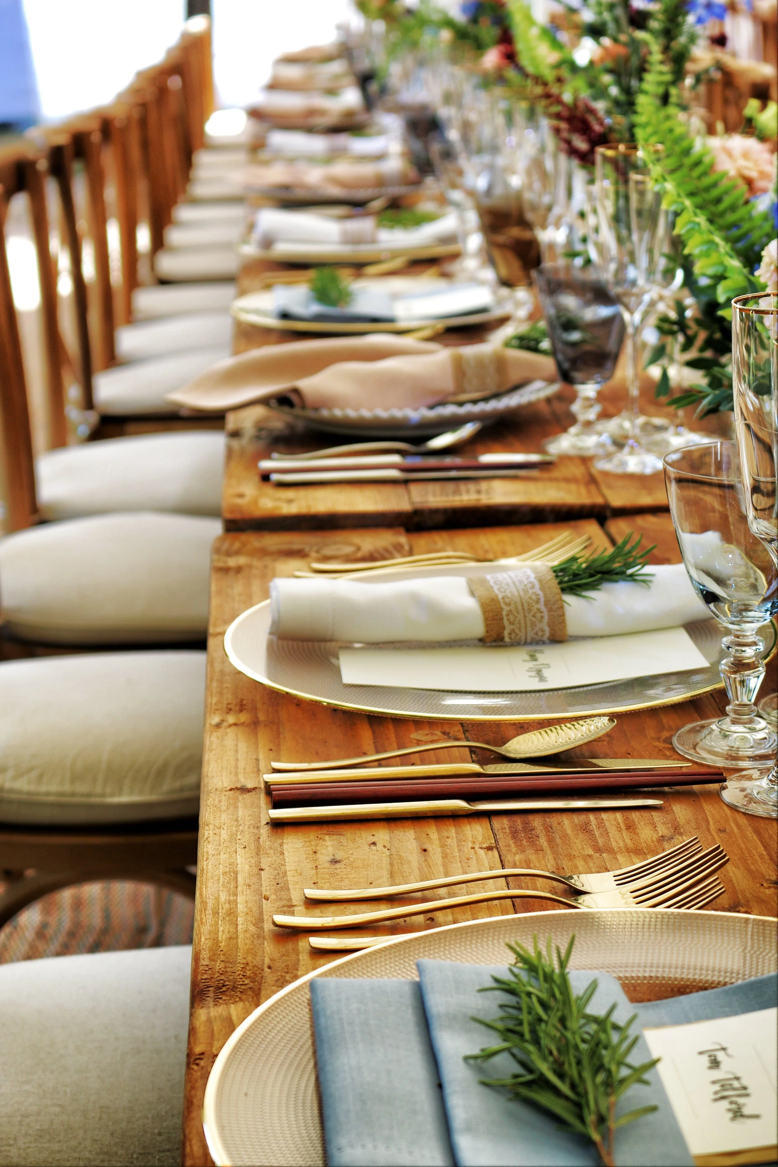 A long wooden dining table set for a formal meal with plates, silverware, glasses, napkins, and decorative greenery, with cream-colored chairs around it.
