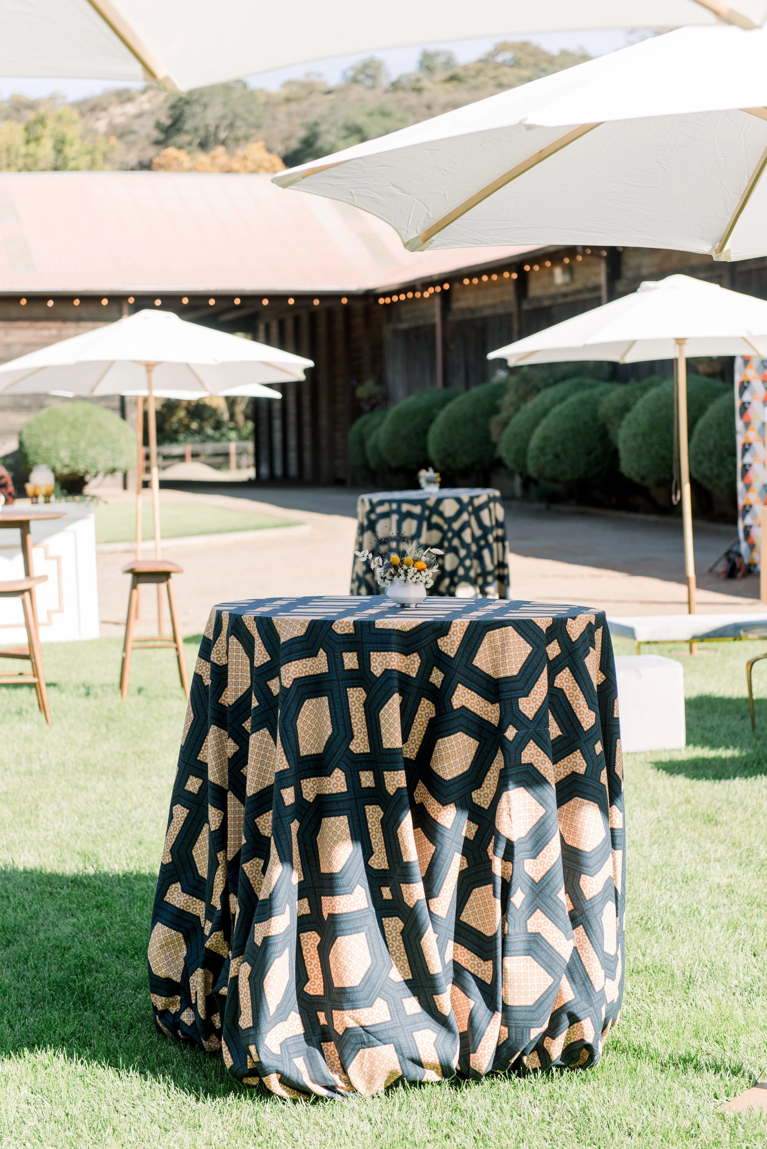 Outdoor event with round tables covered in patterned tablecloths, decorated with small flower arrangements, shaded by large white umbrellas, on a grassy area with a rustic building and green bushes in the background.