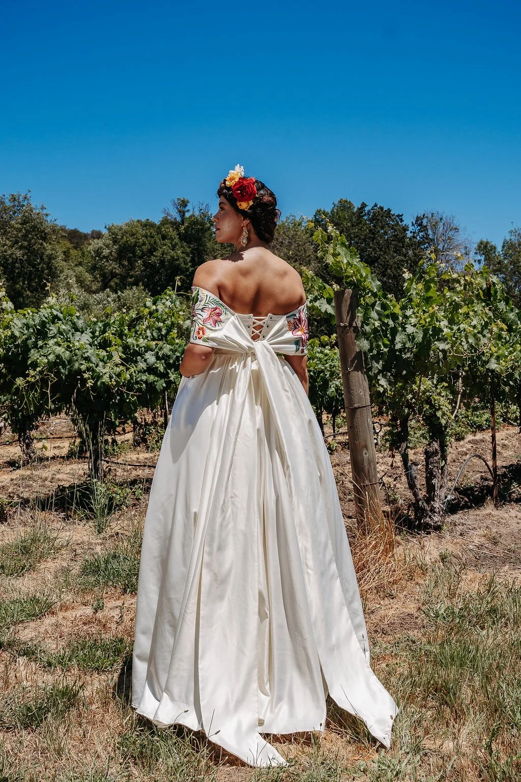 Woman in an off-shoulder white gown with floral embroidery, standing in a vineyard under a clear blue sky, wearing a floral headpiece.