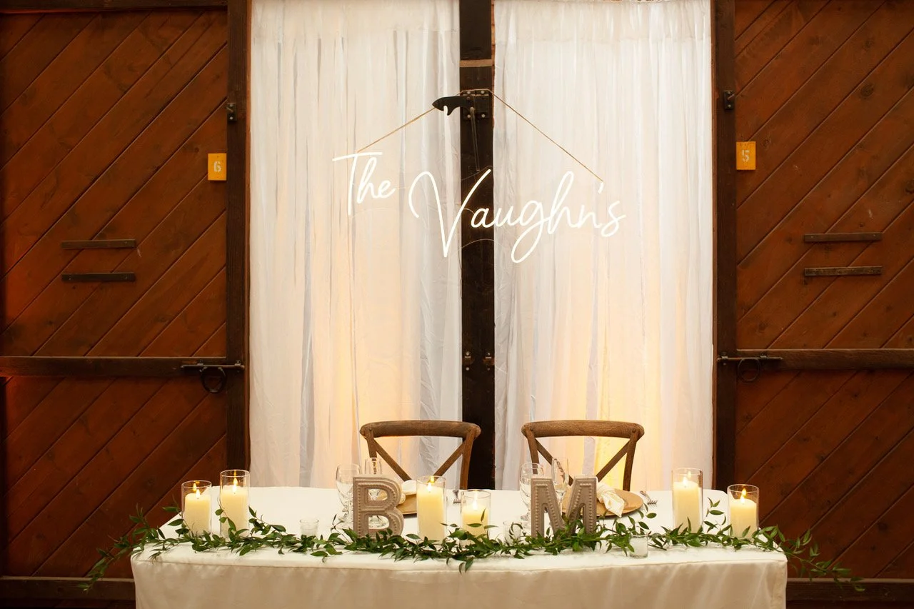 A decorated wedding reception table with candles and greenery, a wooden sign spelling 'BM,' and a neon sign that says 'The Vaughns' in the background.