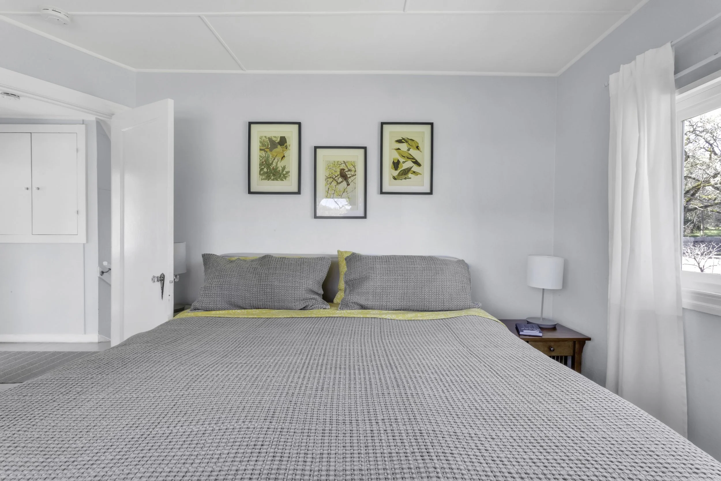 A neatly made bed with gray and yellow bedding, a wooden nightstand with a white lamp and a phone, a window with white curtains, and three framed bird illustrations on the wall in a bright bedroom.