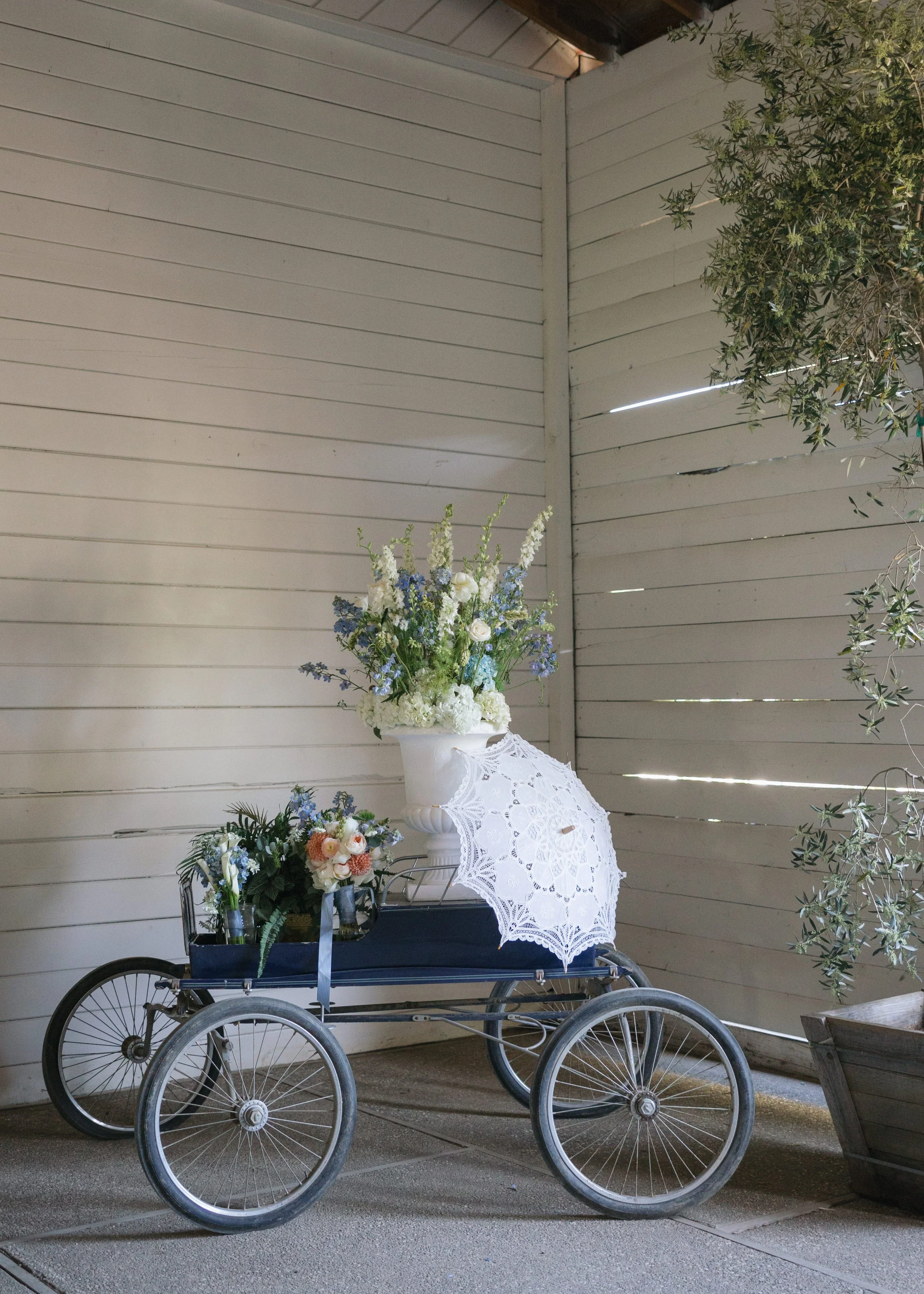 Vintage wheelchair decorated with flowers, a white lace parasol, and greenery, set against a white wooden wall.