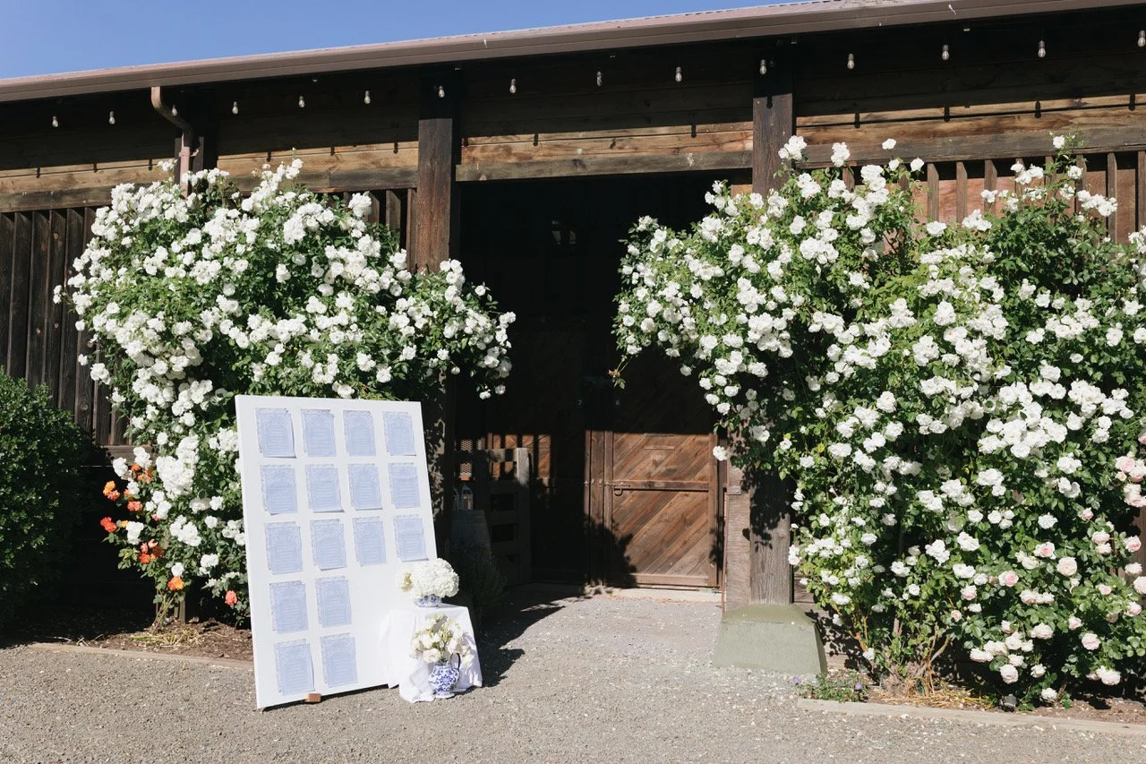 A rustic wooden barn with white blooming bushes flanking the entrance and a display board with multiple white sheets or cards attached, decorated with white flowers in front.