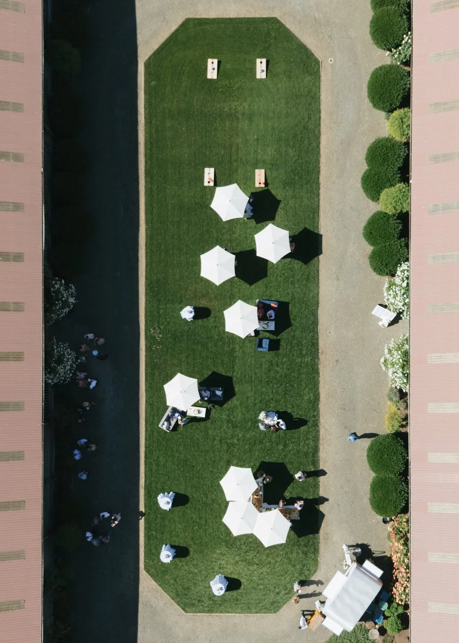 Overhead view of outdoor event with white umbrellas, tables, and people on a grassy area between two buildings with trees and flower bushes.