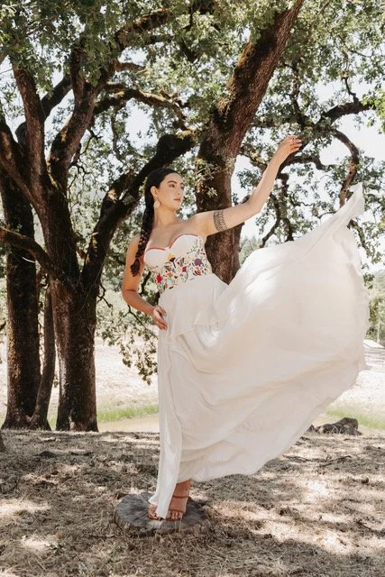 A woman in a white dress with floral embroidery stands outdoors under a large tree, lifting her right leg and holding her dress up slightly, with sunlight filtering through the leaves.