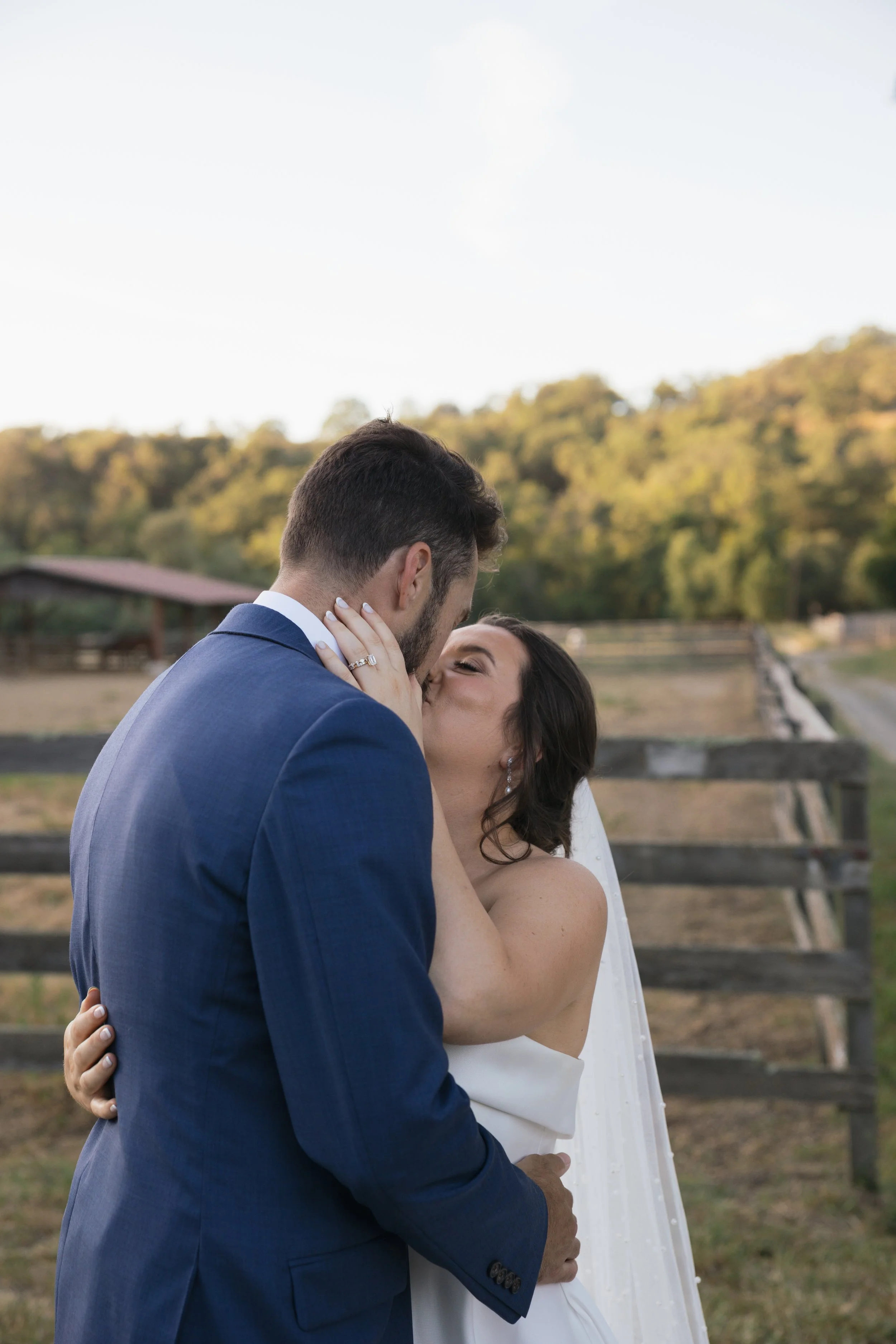 A newlywed couple sharing a kiss outdoors, with the bride in a strapless white dress and the groom in a navy blue suit, against a rural background with trees and a wooden fence.