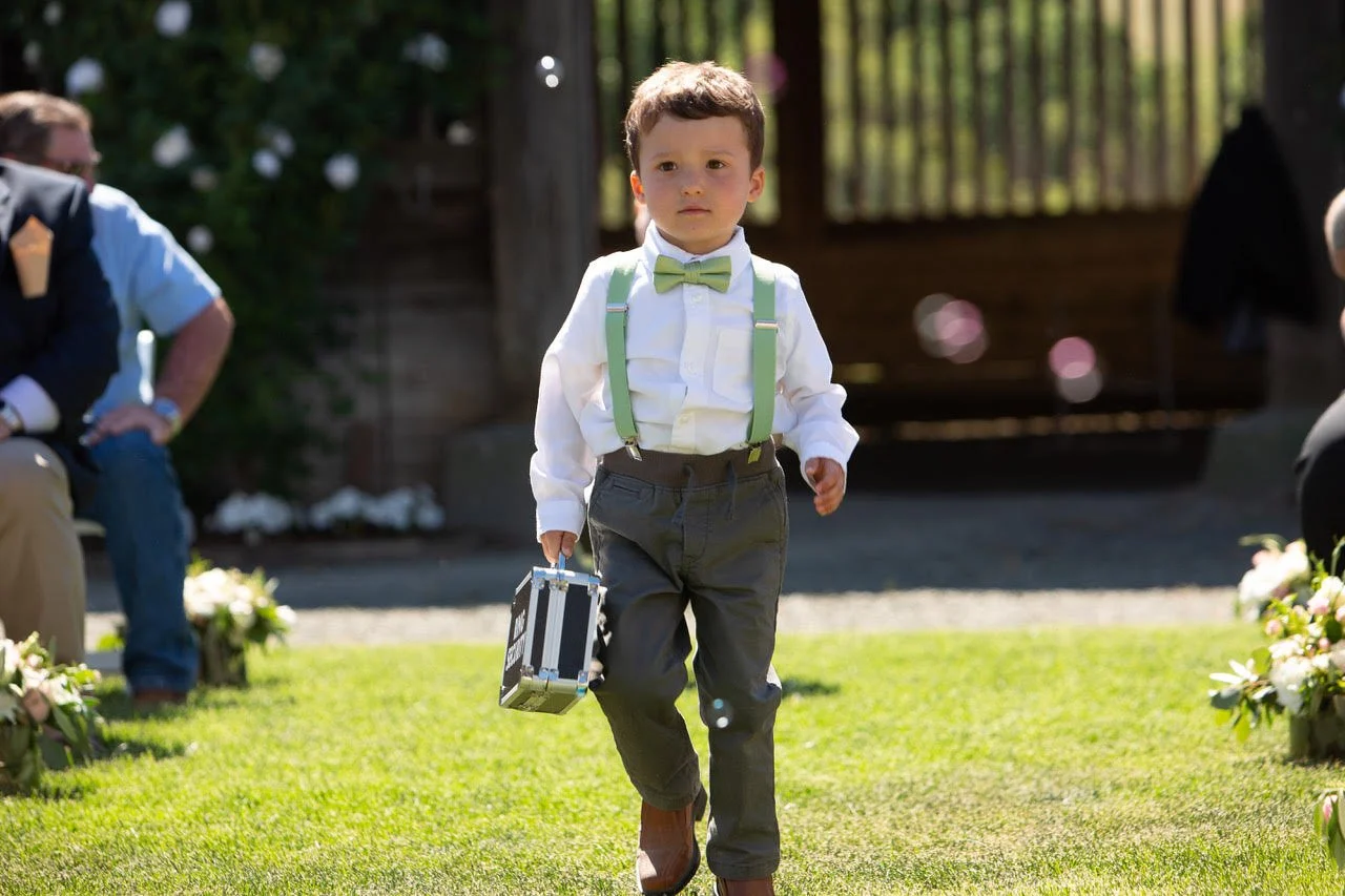 A young boy dressed in formal clothes with a bow tie and suspenders walking on grass, carrying a small suitcase.