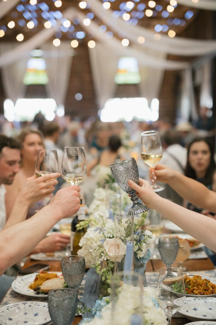 People at a wedding reception raising glasses of white wine for a toast in a decorated barn with string lights and floral centerpieces.