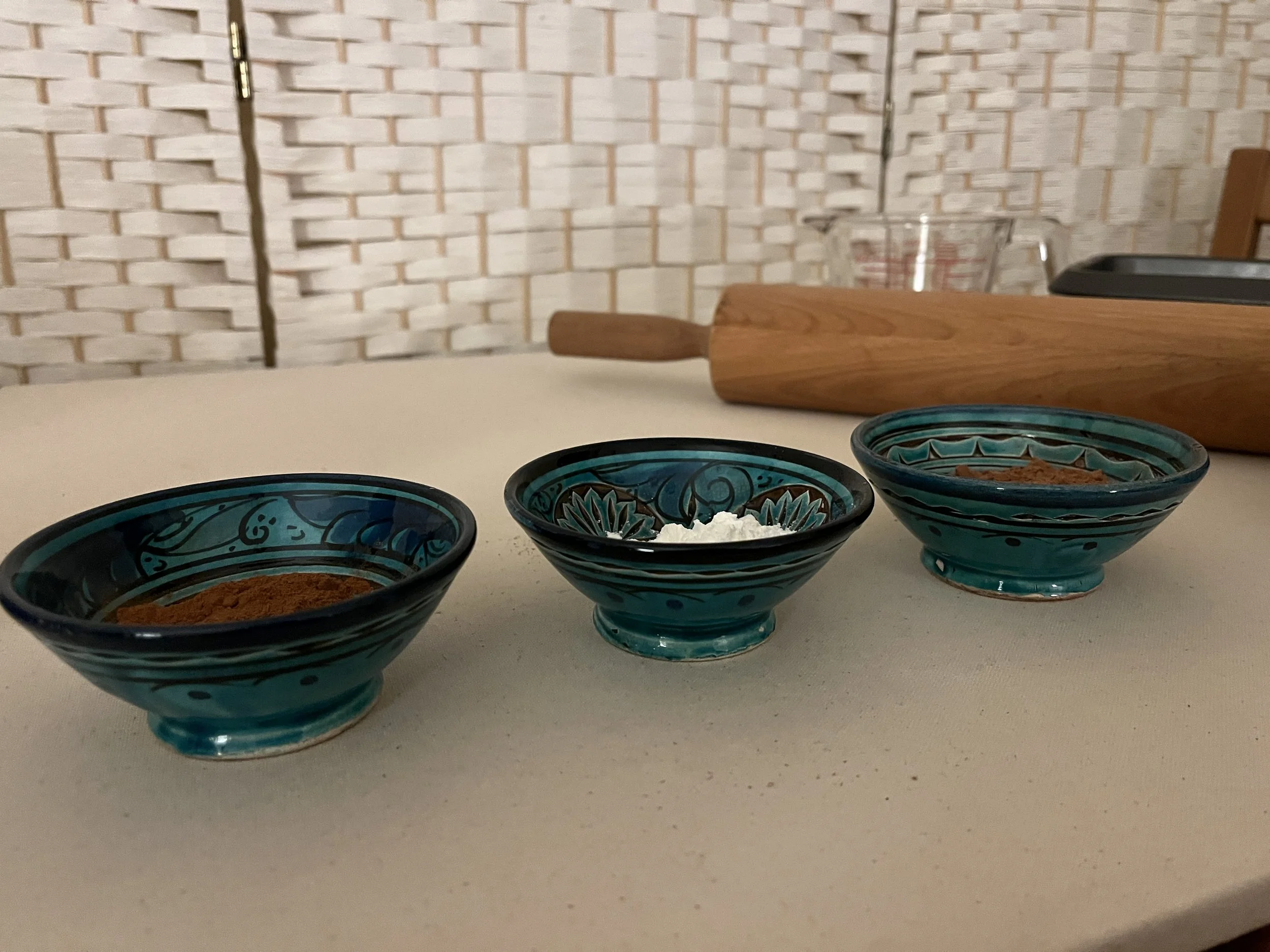 Three small turquoise blue and navy blue moroccan bowls on a beige background filled with spices and baking powder with a rolling pin, liquid measuring cup and bread pan in the right corner of the photo. The vertical plane behind is white wicker.