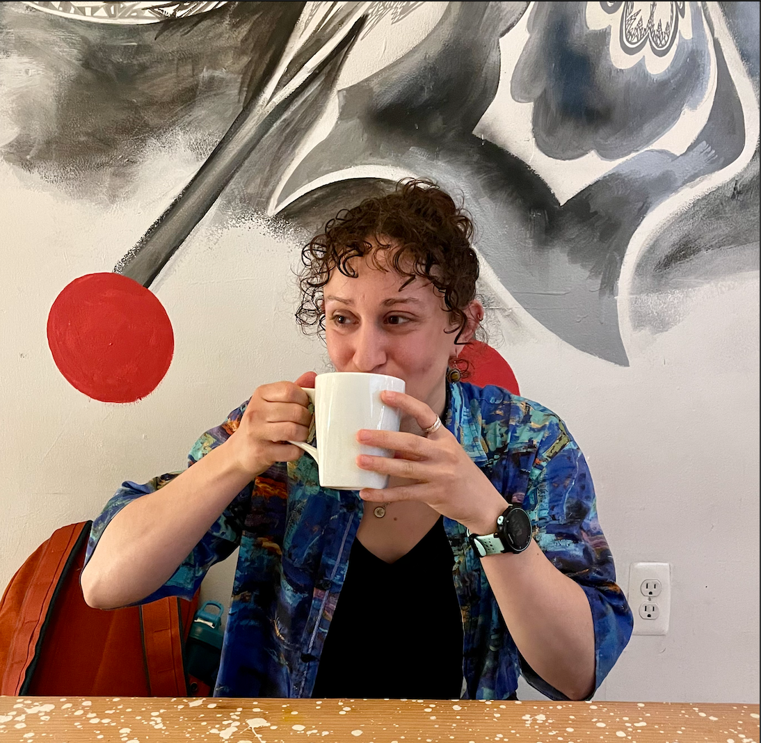 A woman with brown curly hair, wearing a blue patterned jacket, sitting at a table with a mug in her hands, drinking. Behind her is a mural with abstract black, white, and red shapes.
