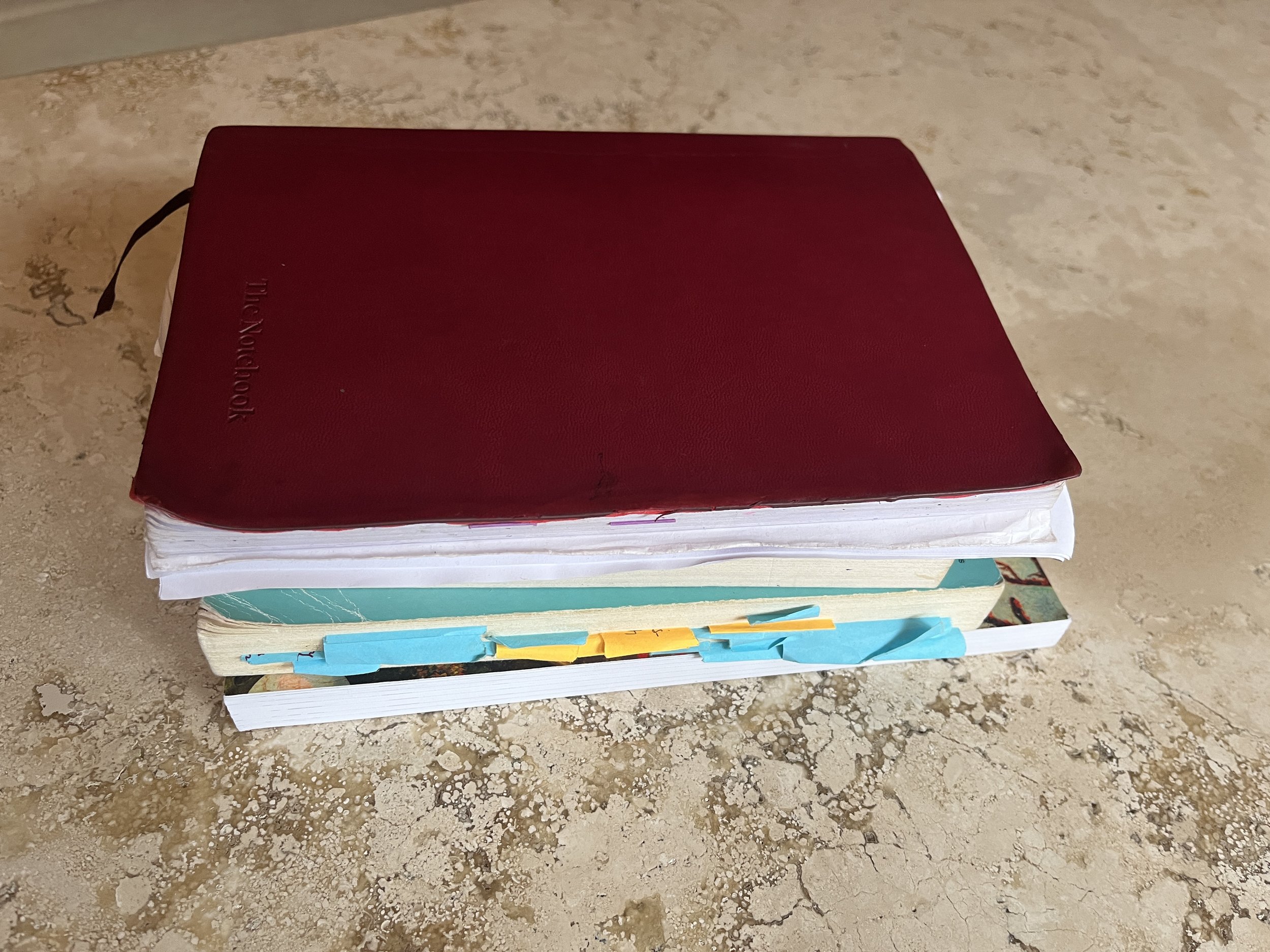 A stack of four books with the top book being cranberry colored and the third book filled with orange and blue post-its against a beige marble background.