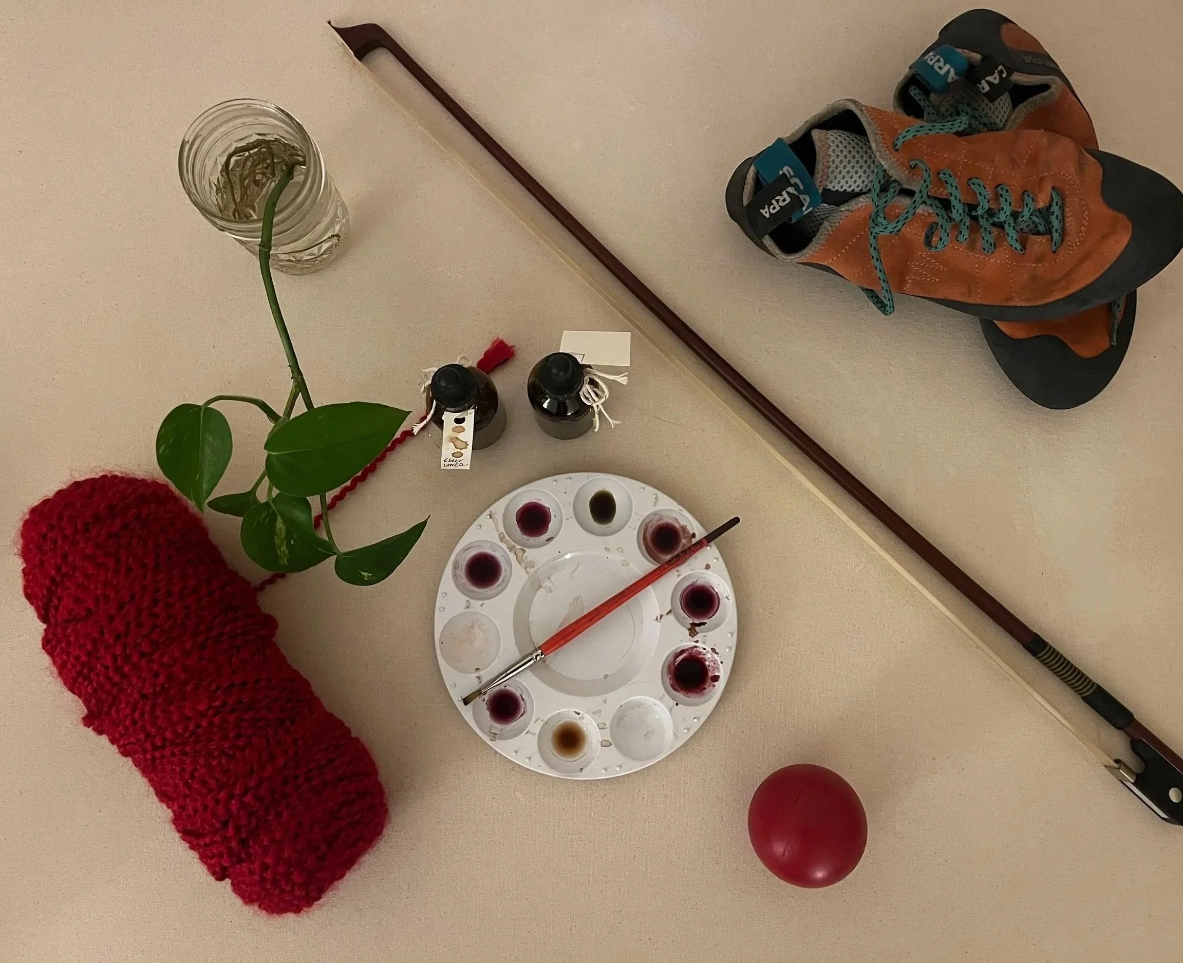 a birds-eye view of red yarn, a pothos plant propagating in a water glass, two dropper bottles of ink, a palatte of paint and paint brushes, a bow for a string instrument, and orange climbling shones, which all sit on top of a beige canvas.