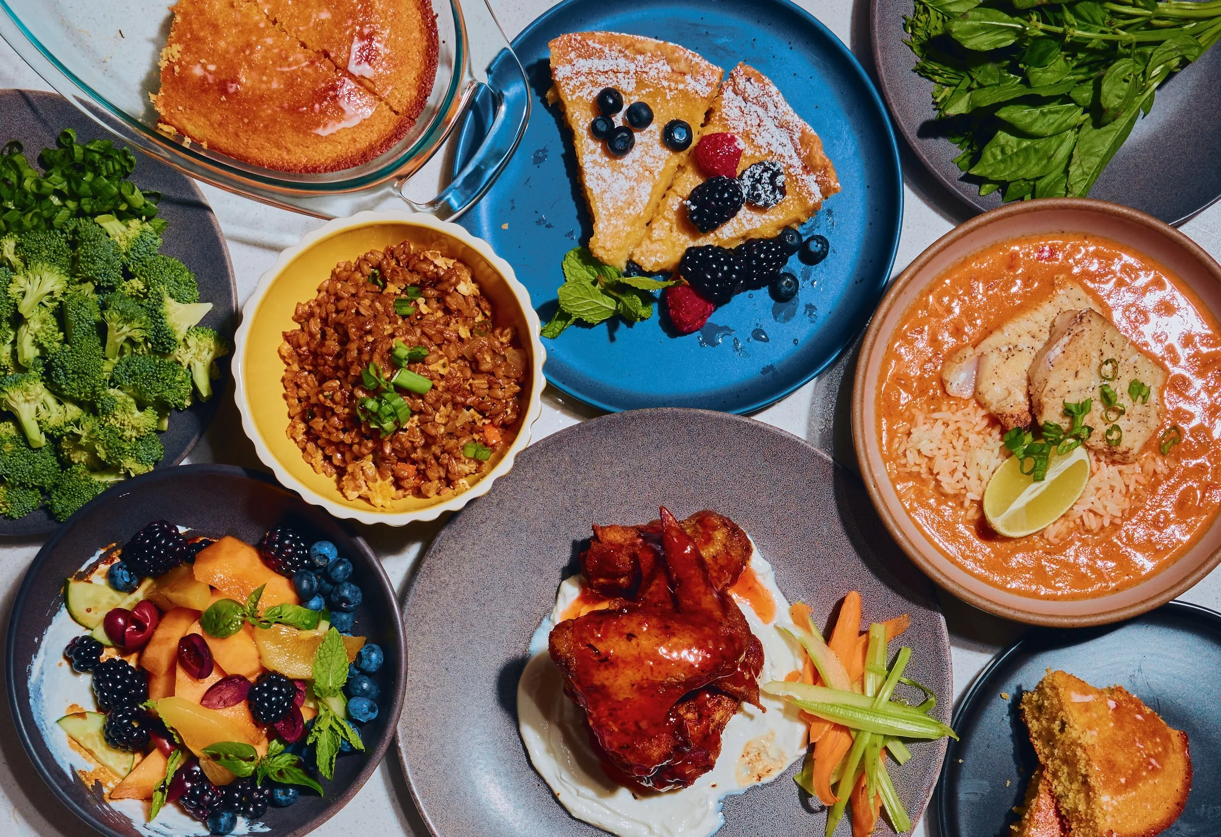 A variety of dishes on a table, including broccoli, a fruit salad, two slices of cake with berries, a bowl of rice and chicken in sauce, a plate with roasted chicken and vegetables, a plate of plantains, and a bowl of lentils.