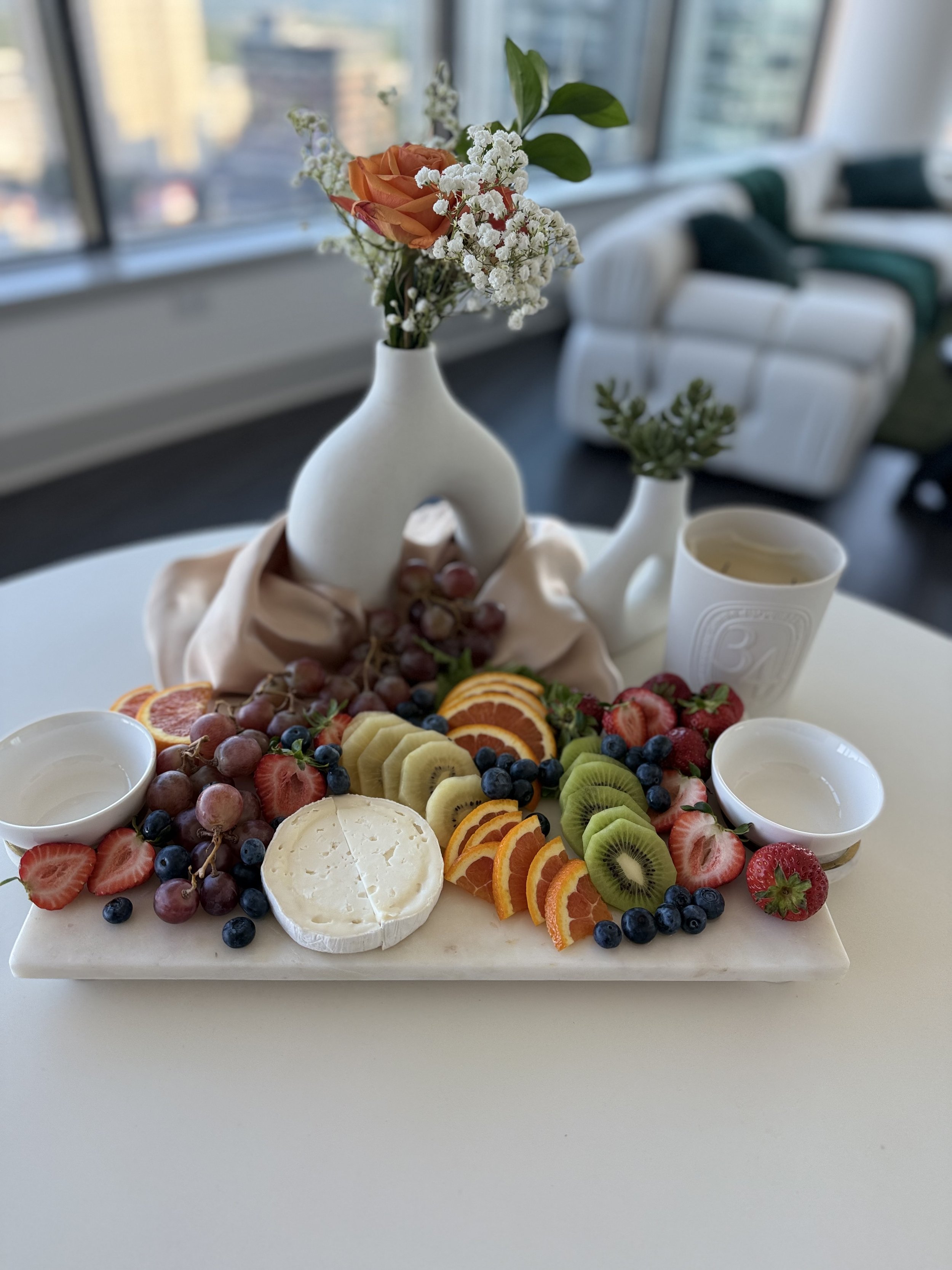 A marble platter with assorted sliced fruits, cheese, and berries, with a white vase holding a flower arrangement, set on a white table, in a room with large windows and modern furniture.