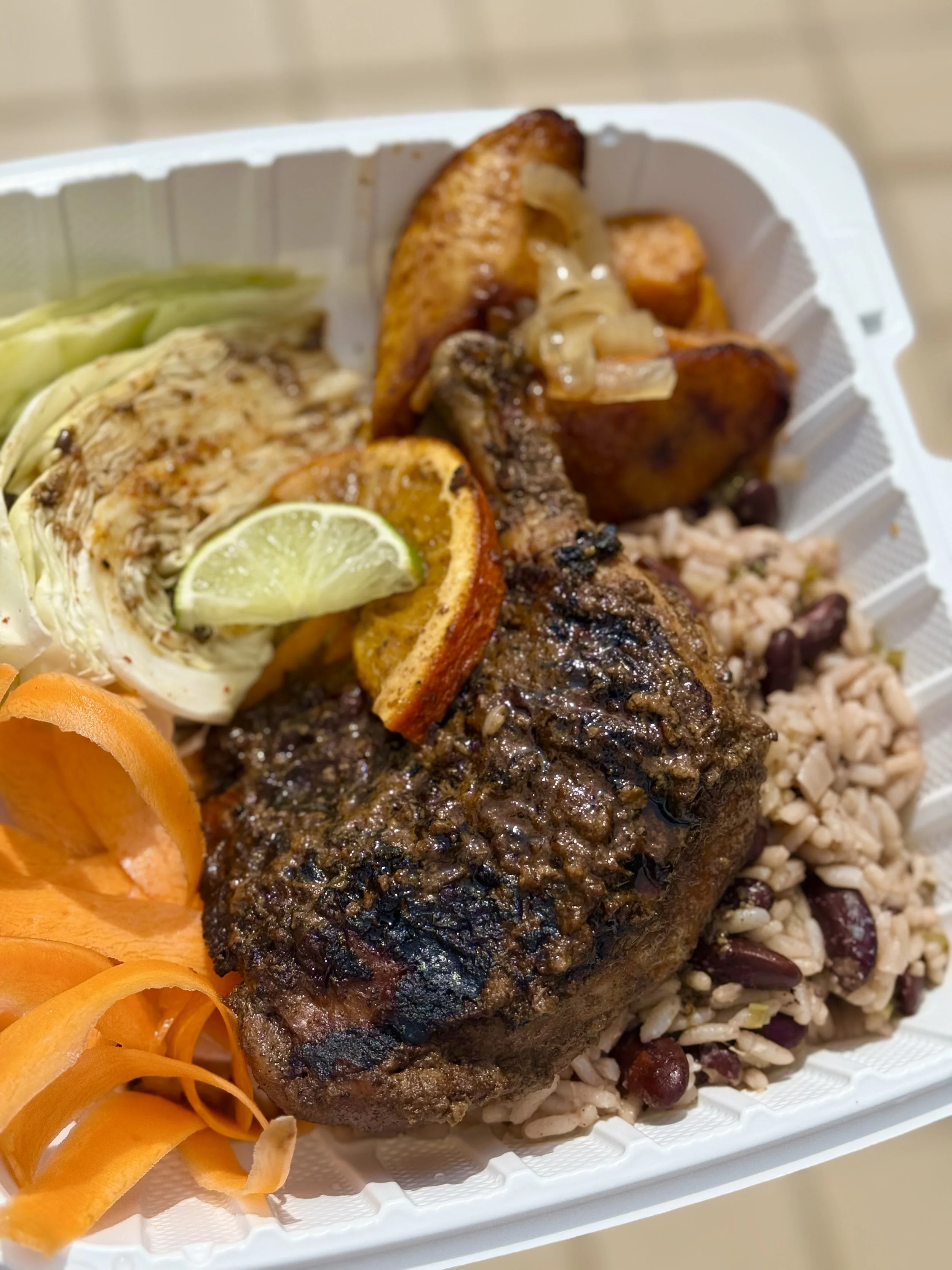 Close-up of a takeout food tray with grilled meat, rice and beans, roasted plantains, shredded lettuce, sliced lime, and a vegetable garnish.