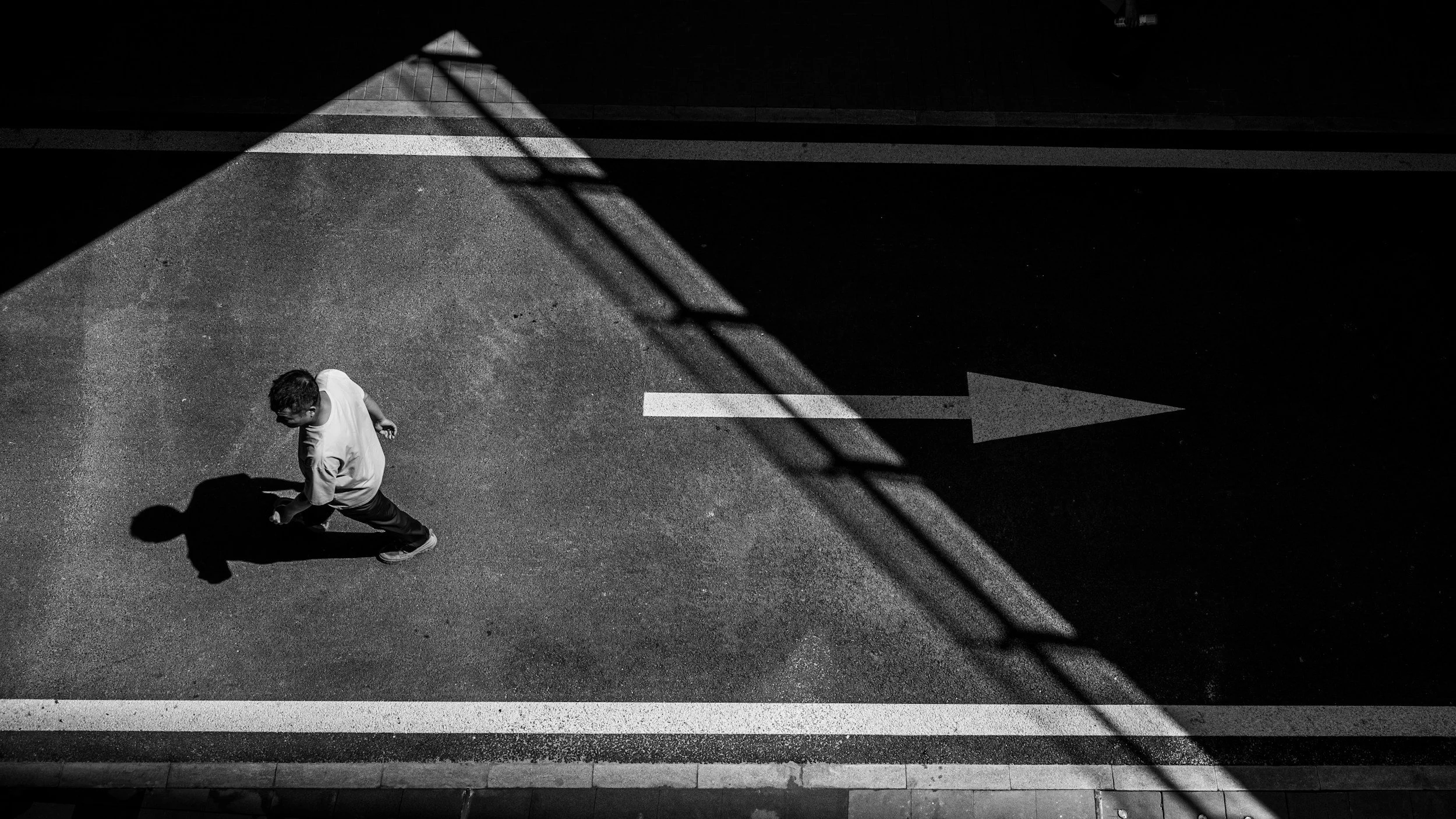 A man walking along a street is casting a shadow on the pavement, which appears darker and more shaded due to a shadow from a nearby structure. The street has a right-pointing arrow painted on it, and a white line marks the curb.