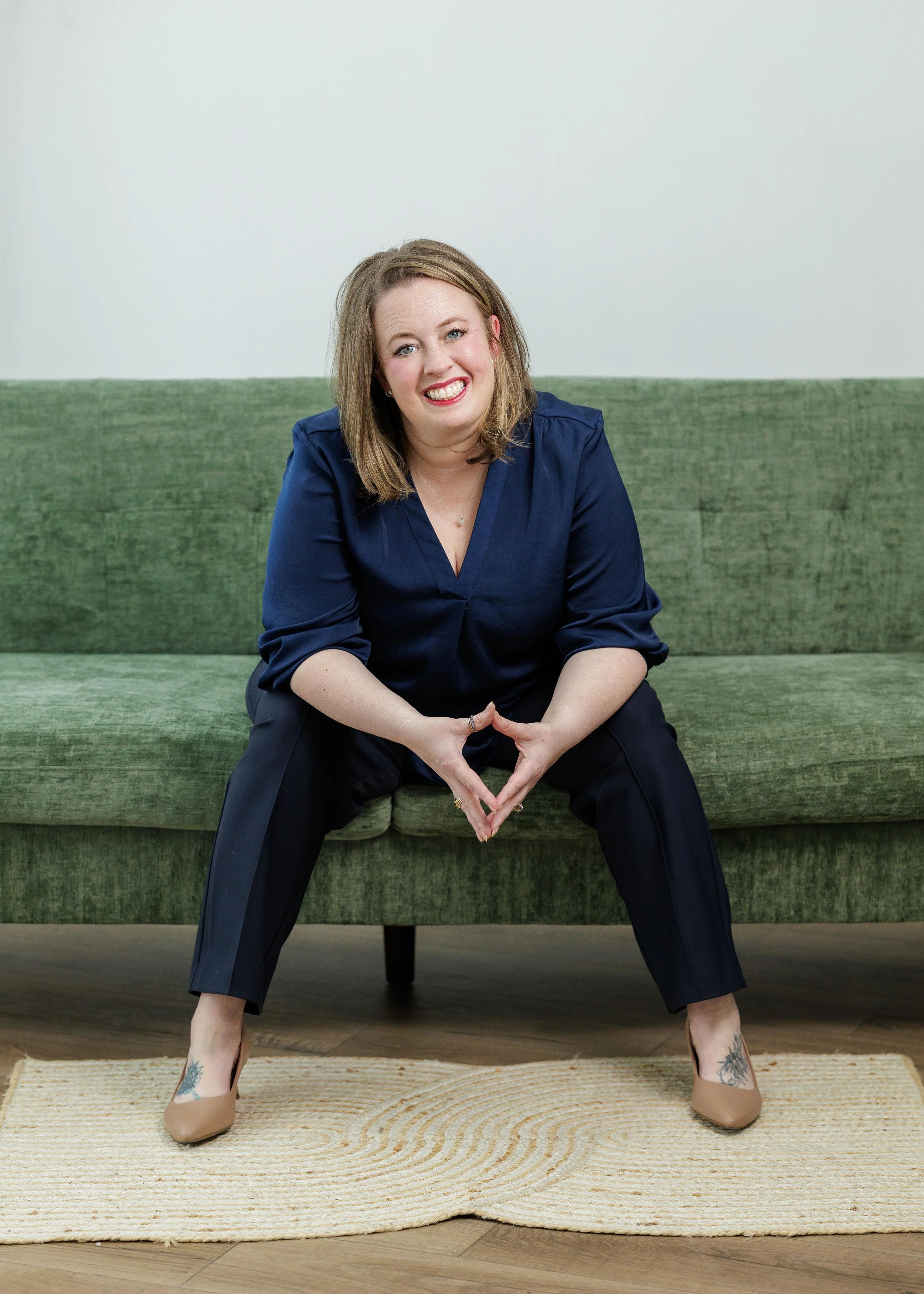 A woman with blonde hair and tattoos on her ankles, sitting on a green sofa with her legs wide apart, smiling and forming a heart with her hands.