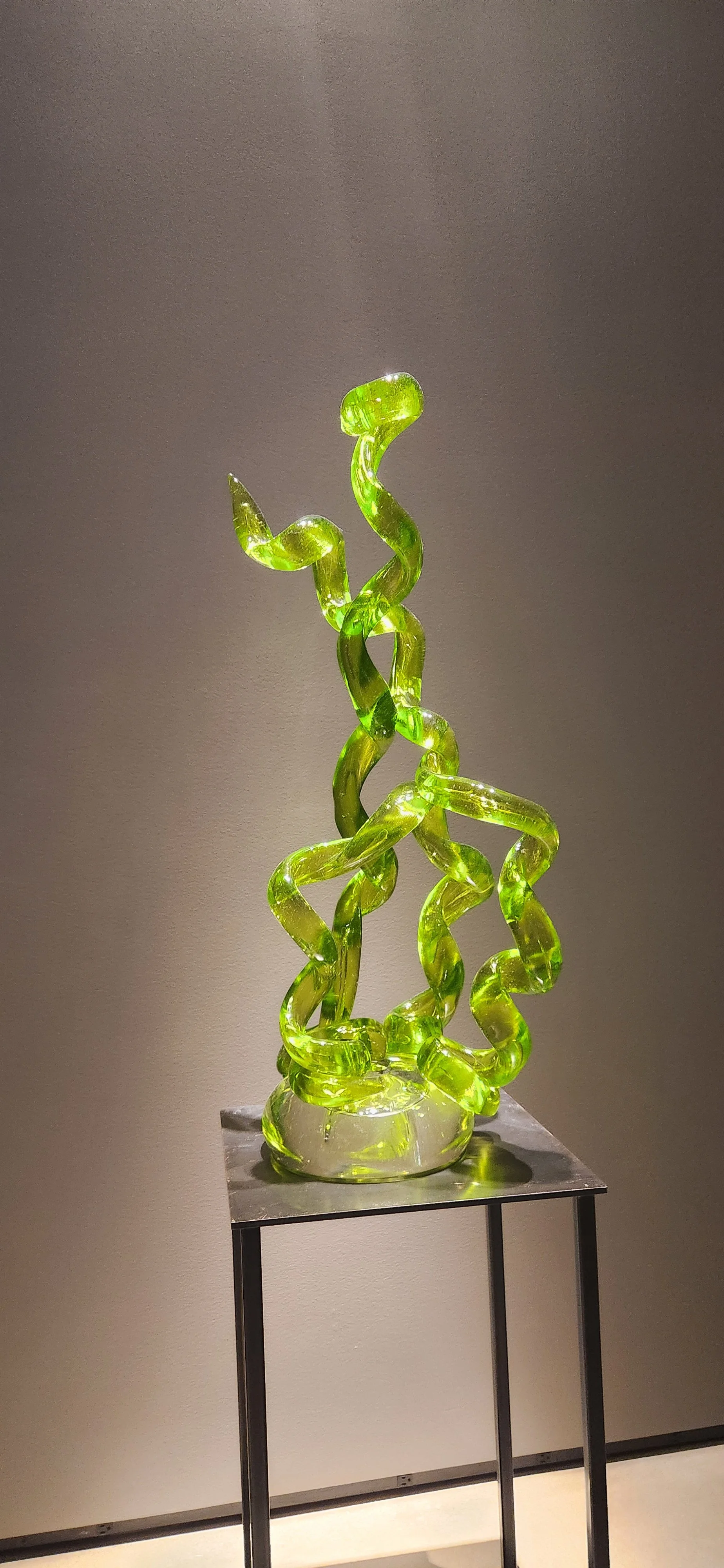 Glass sculpture with intertwined, twisting, lime-green glass tubes displayed on a metal stand.
