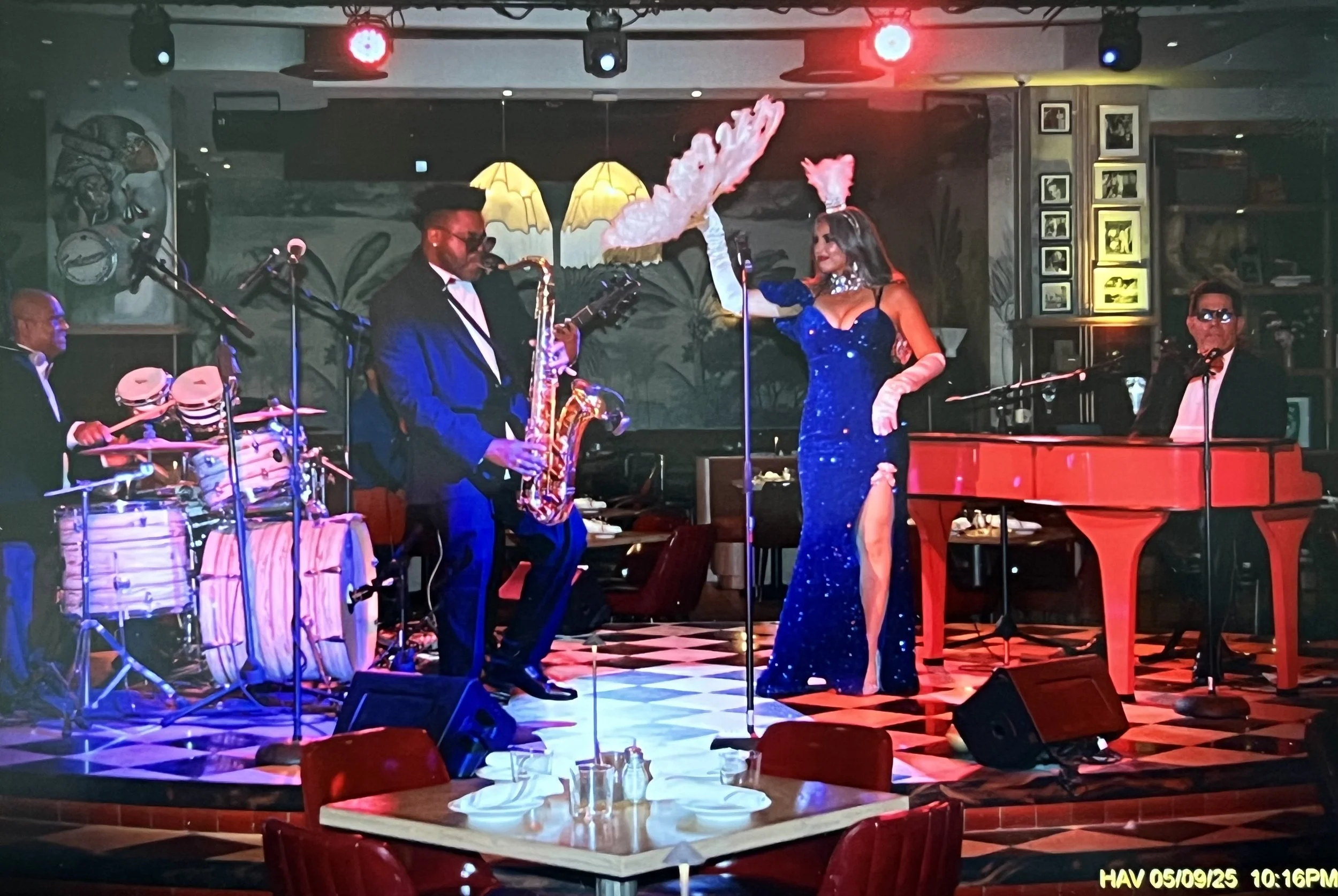 A jazz band performing live on stage in a restaurant, featuring a singer in a sparkly blue gown with a feathered headpiece, a saxophonist, a drummer, and a pianist.