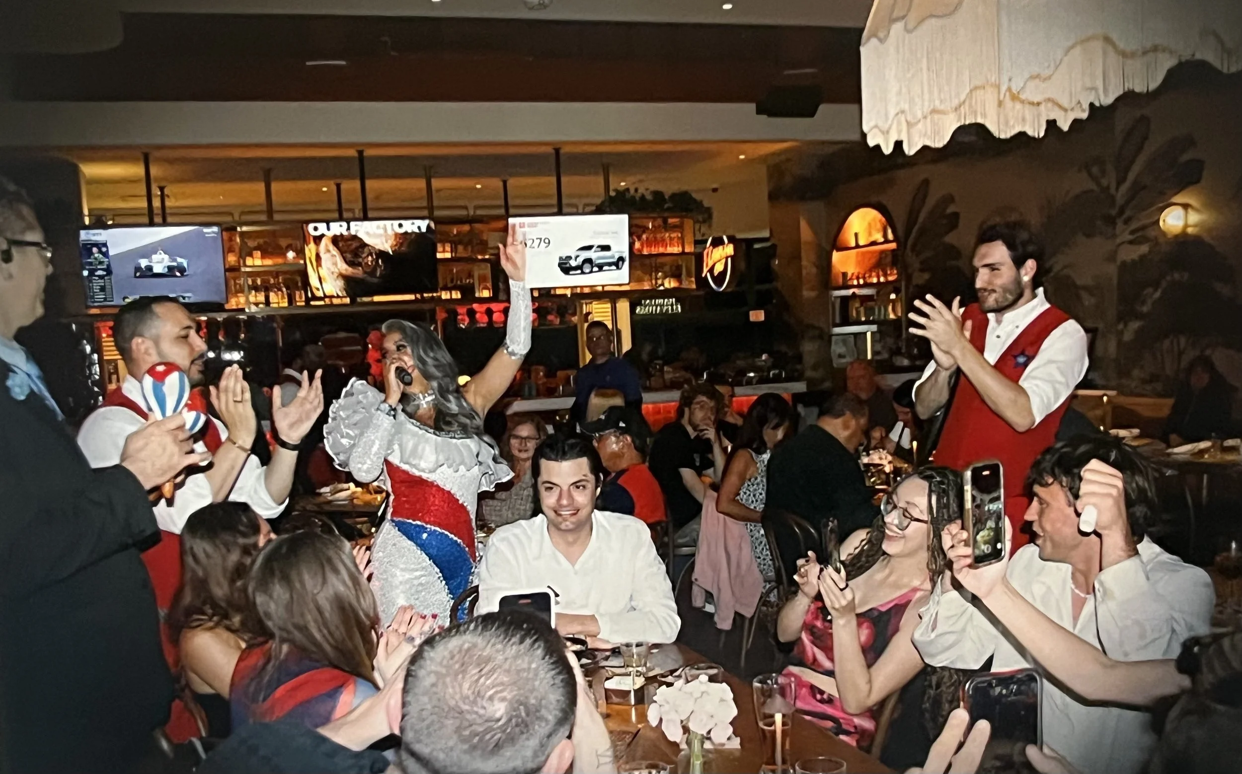People celebrating at a restaurant or bar, with a singer holding a microphone, a woman raising her hand, others clapping and recording, in a lively social gathering with a bar in the background.