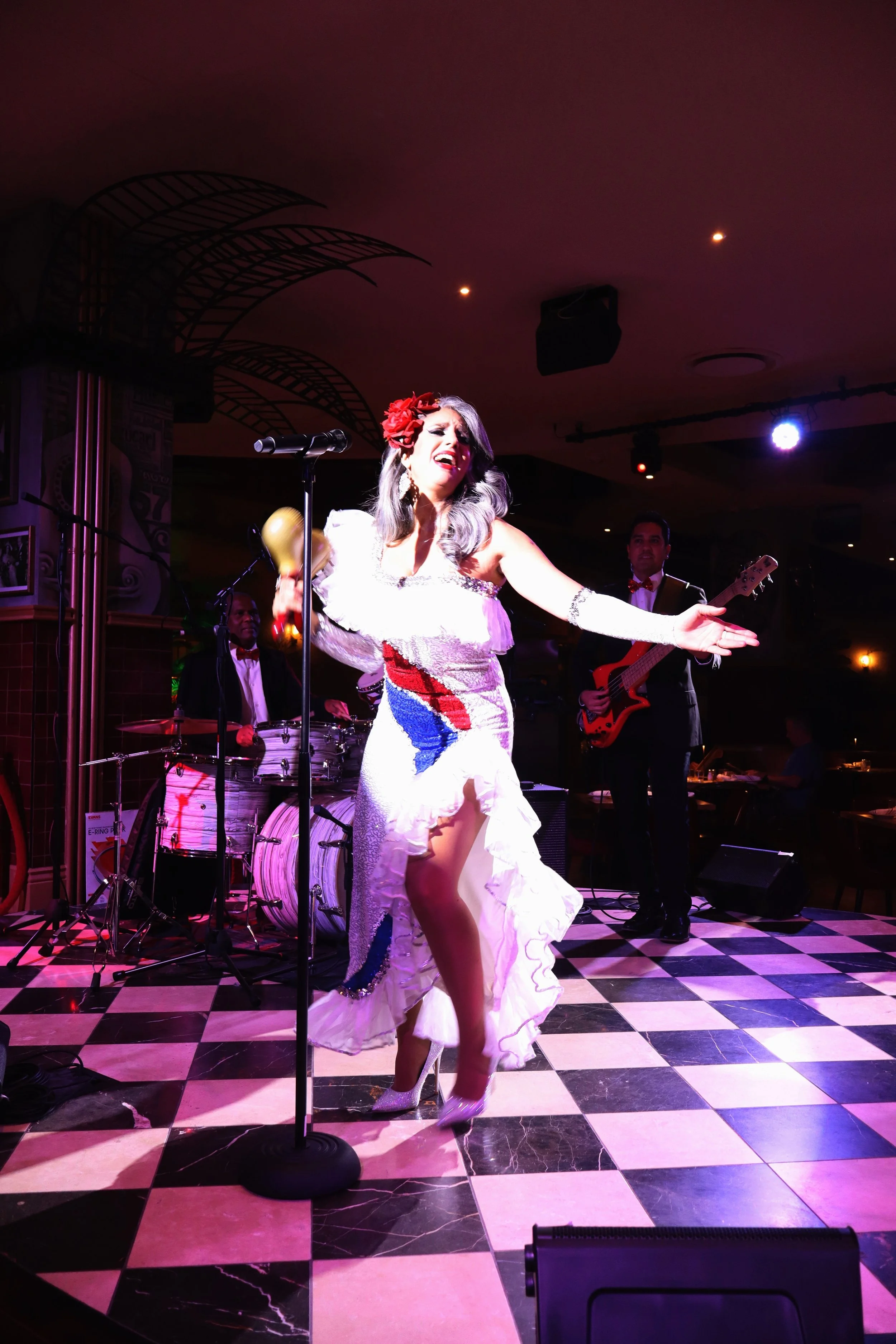 Performer singing on stage with musical band, woman in white dress with ruffled skirt, red flower headpiece, holding a maraca, black and white checkered floor, dim stage lighting.