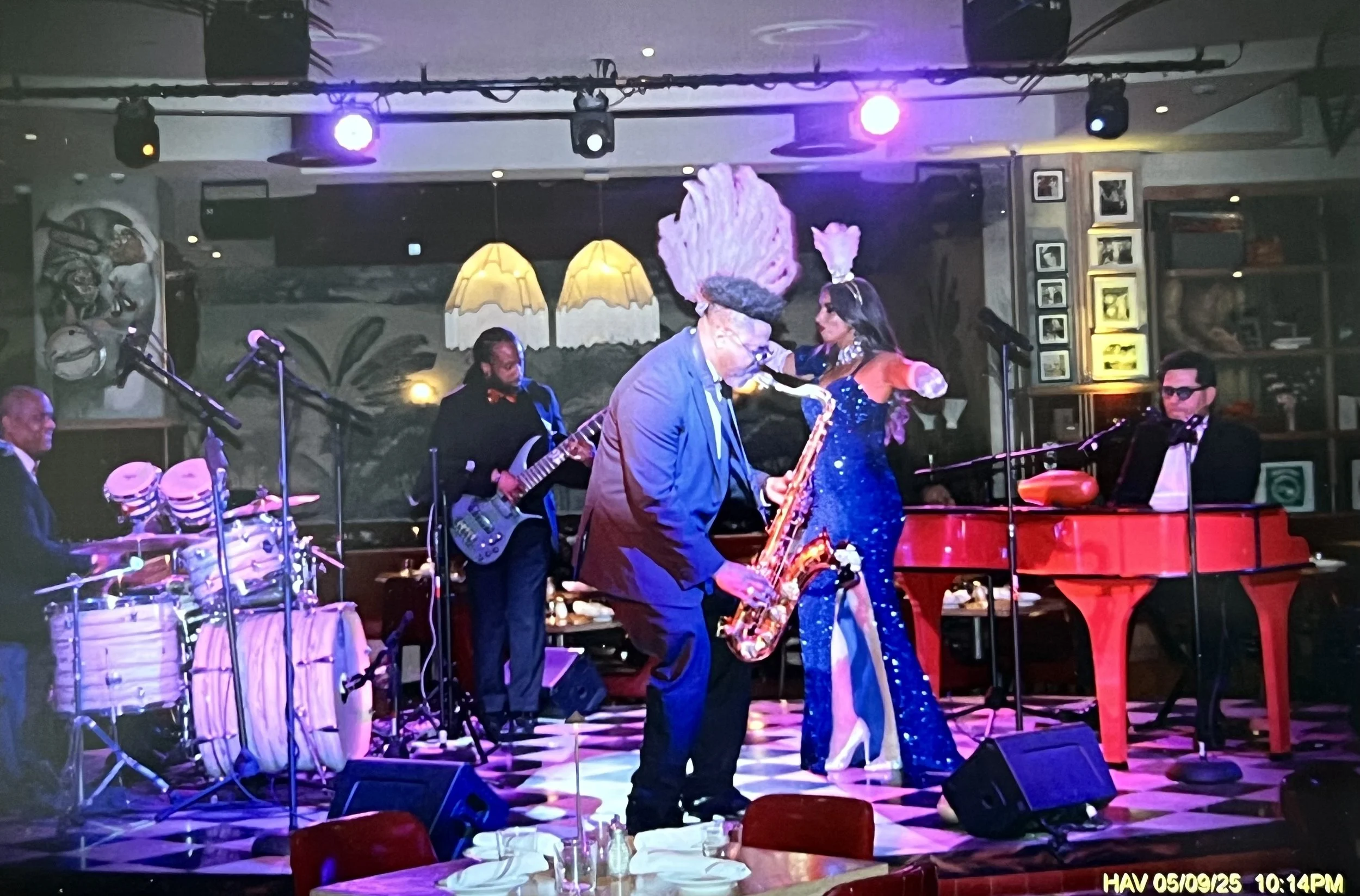 Live jazz band performing at a restaurant, featuring a male saxophonist with a tall, lavender hairstyle, a female singer in a sparkly blue dress, a pianist, a bassist, and a percussionist. The scene is lit with colorful stage lights and has a checker