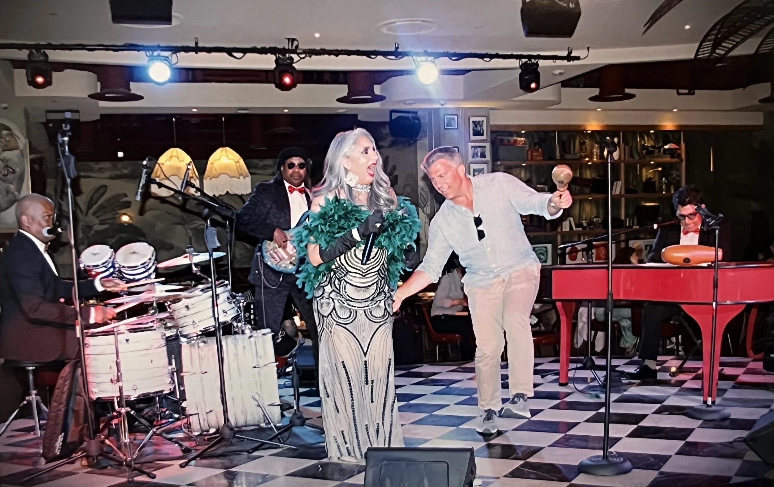A lively musical performance in a cozy venue featuring a group of performers on stage, including a woman in an elaborate dress singing into a microphone, a man smiling and dancing, a drummer, and a keyboard player, with vintage decor and warm lightin