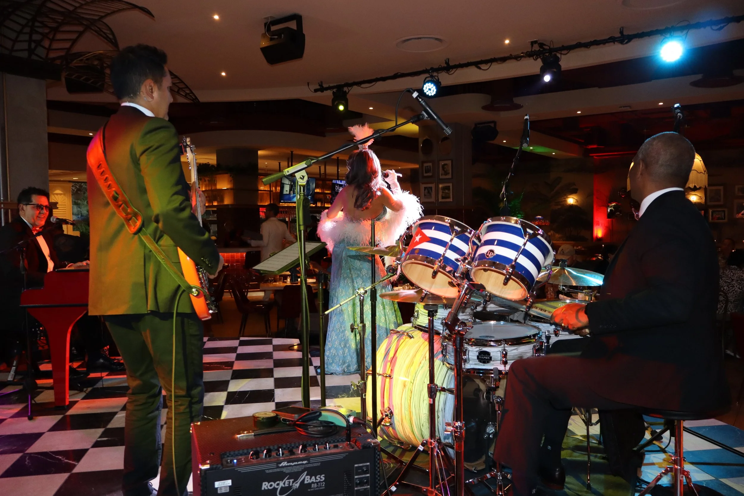 A band performing on stage with a female singer in a long dress, a guitarist, and a drummer, in a dimly lit venue with colorful lighting and checkered floor.