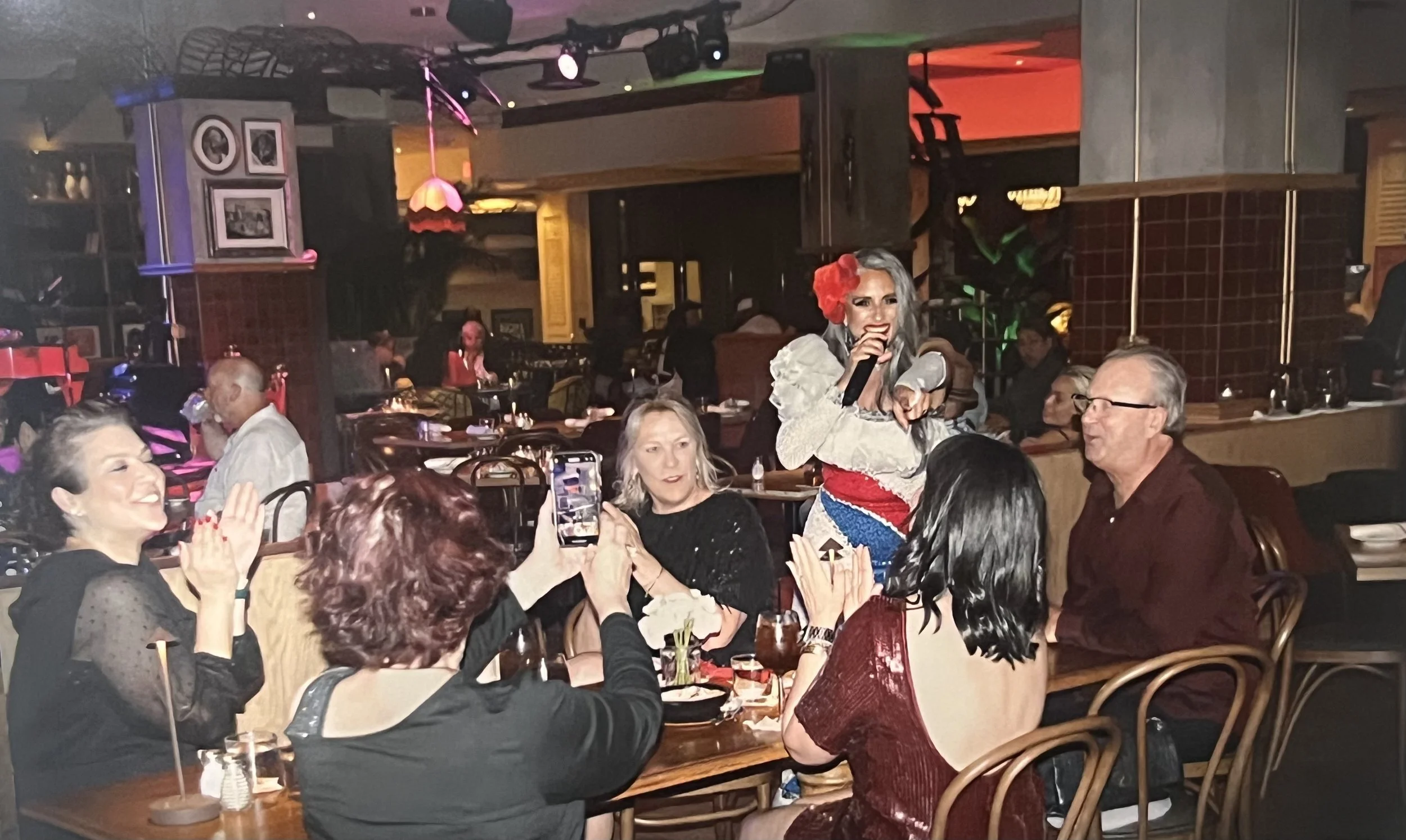 A woman with gray hair, a red flower in her hair, and colorful clothing is singing or talking into a microphone at a restaurant or bar. She is surrounded by seated people clapping, taking photos, and enjoying the performance. The setting has dim ligh