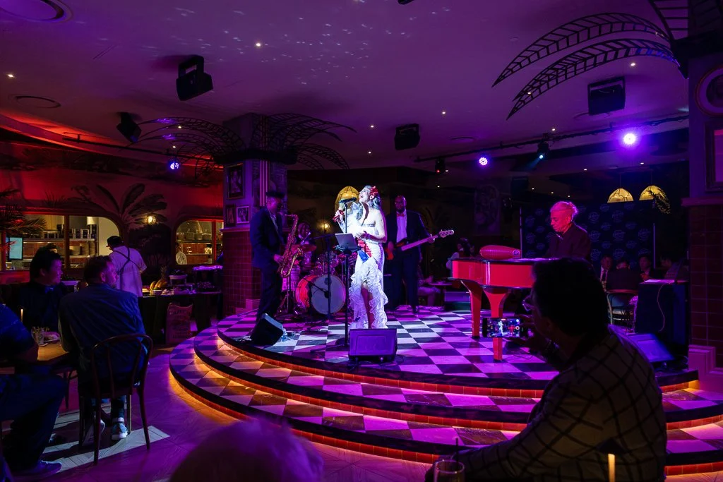 Live jazz band performing on small checkered stage in a dimly lit lounge with purple and red lighting, surrounded by seated audience.