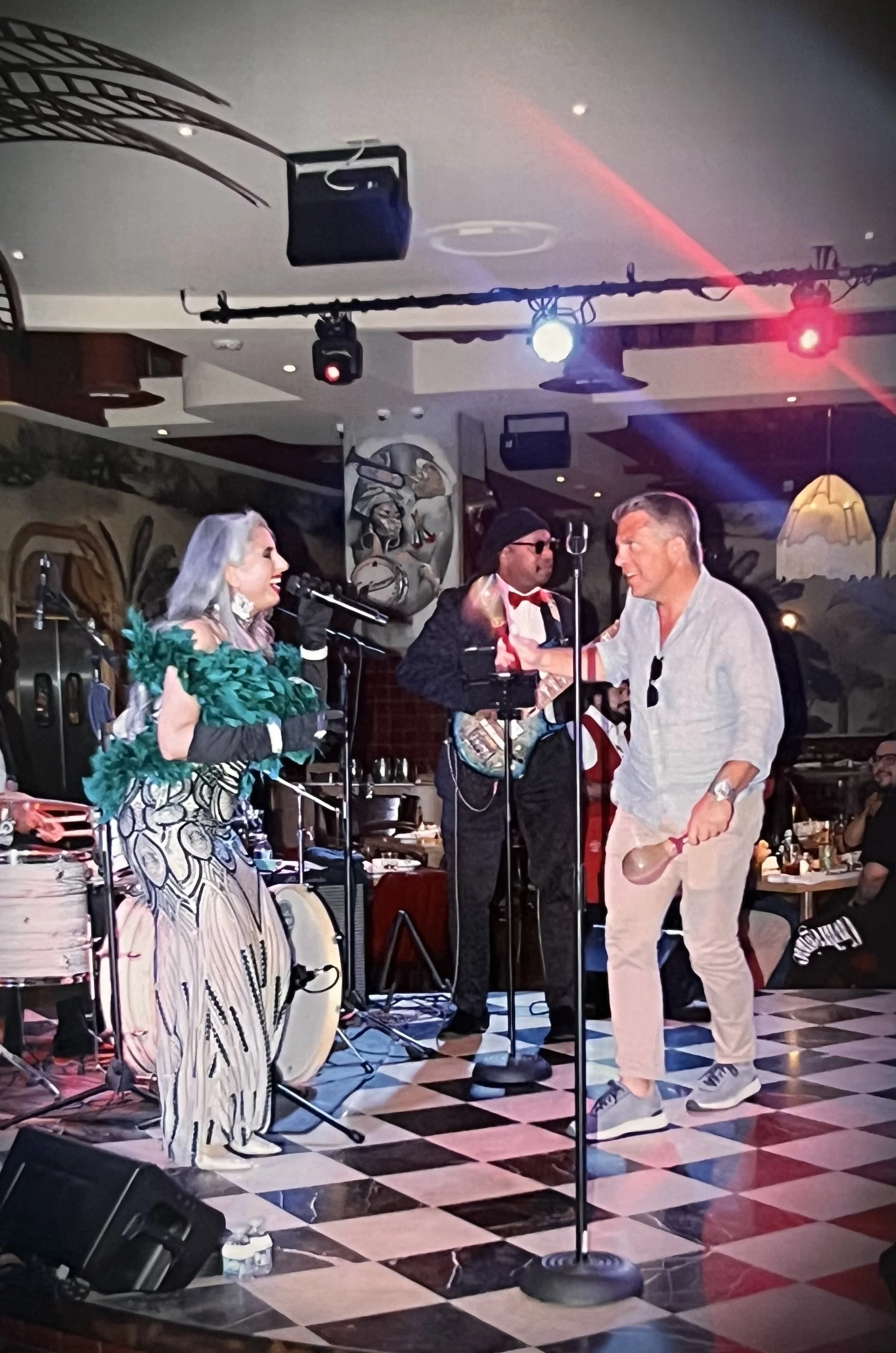 Performance on stage with three musicians, including a woman with gray hair wearing a vibrant green feathered top, a man with sunglasses and a bow tie, and another man with a musical instrument, in a lively indoor venue with dancing and audience memb