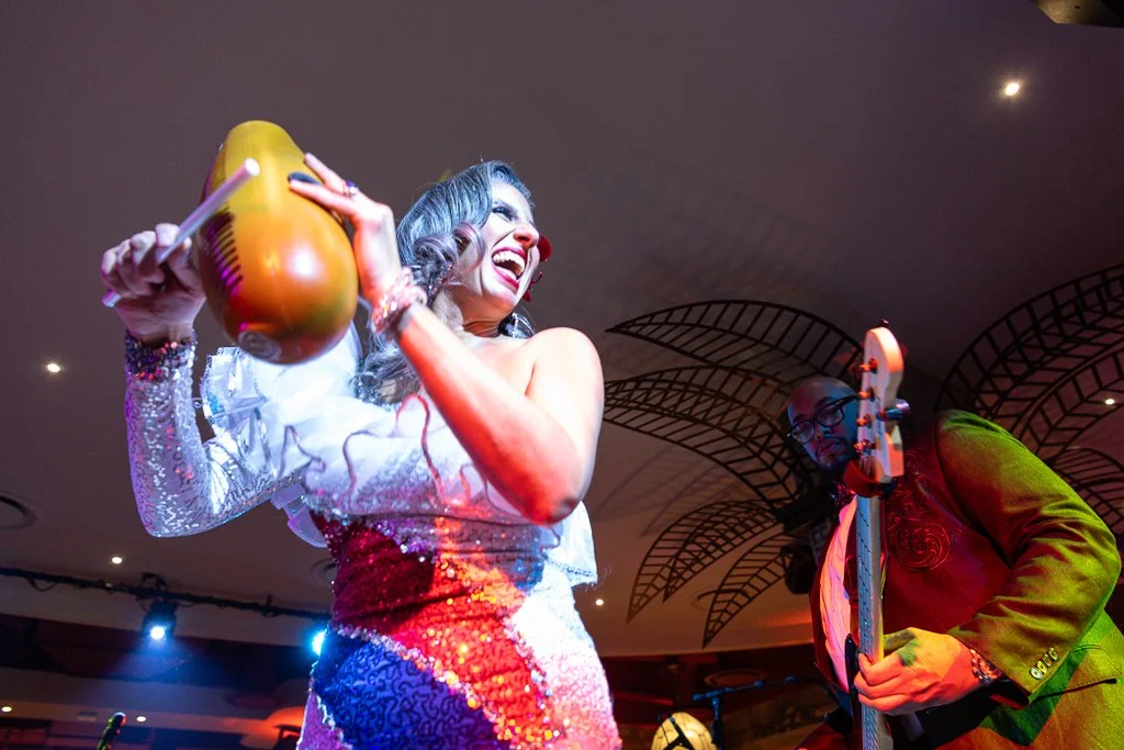 Woman in a glittering outfit playing a guitar with a musician in a colorful suit playing a bass guitar behind her at an indoor event.