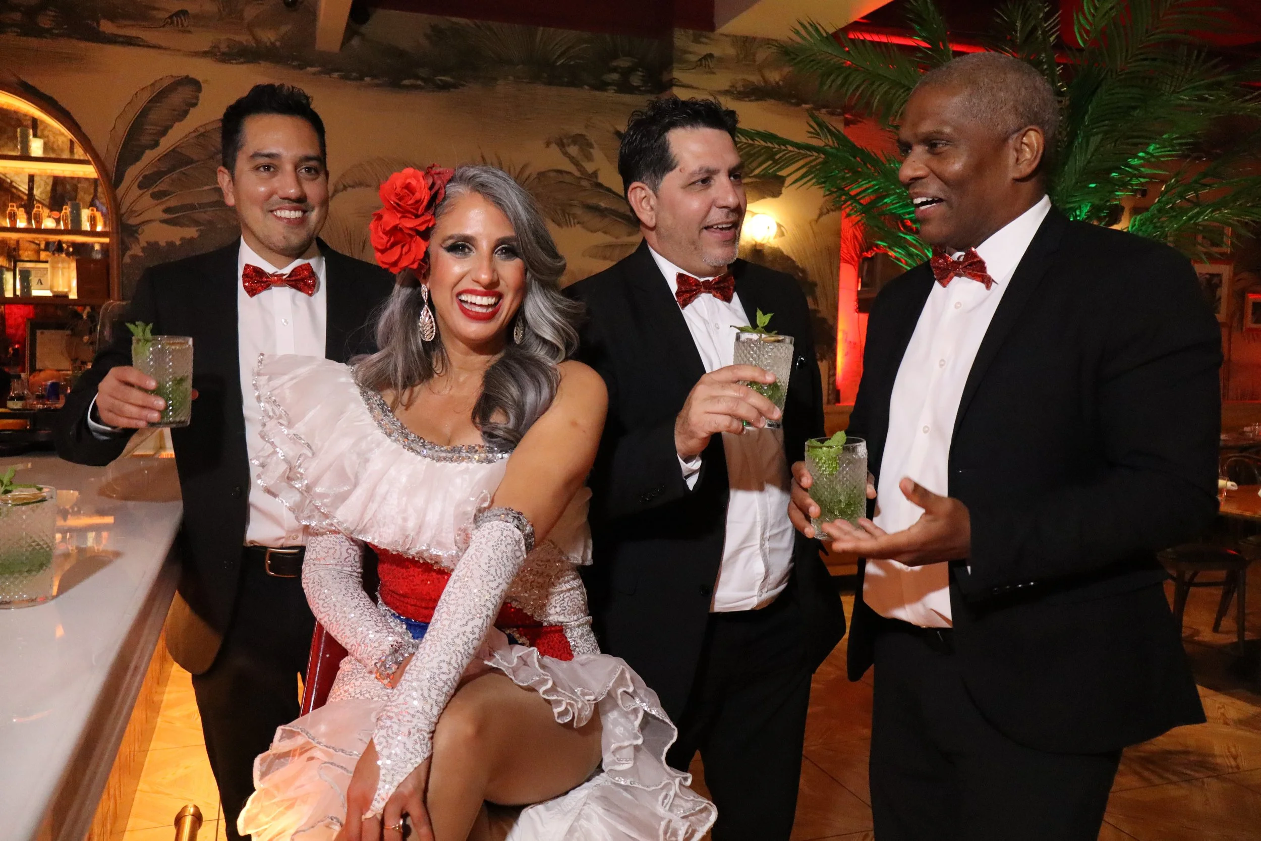 Four people dressed in formal attire celebrating at a party, with drinks in hand, in a warmly lit room with tropical decor.